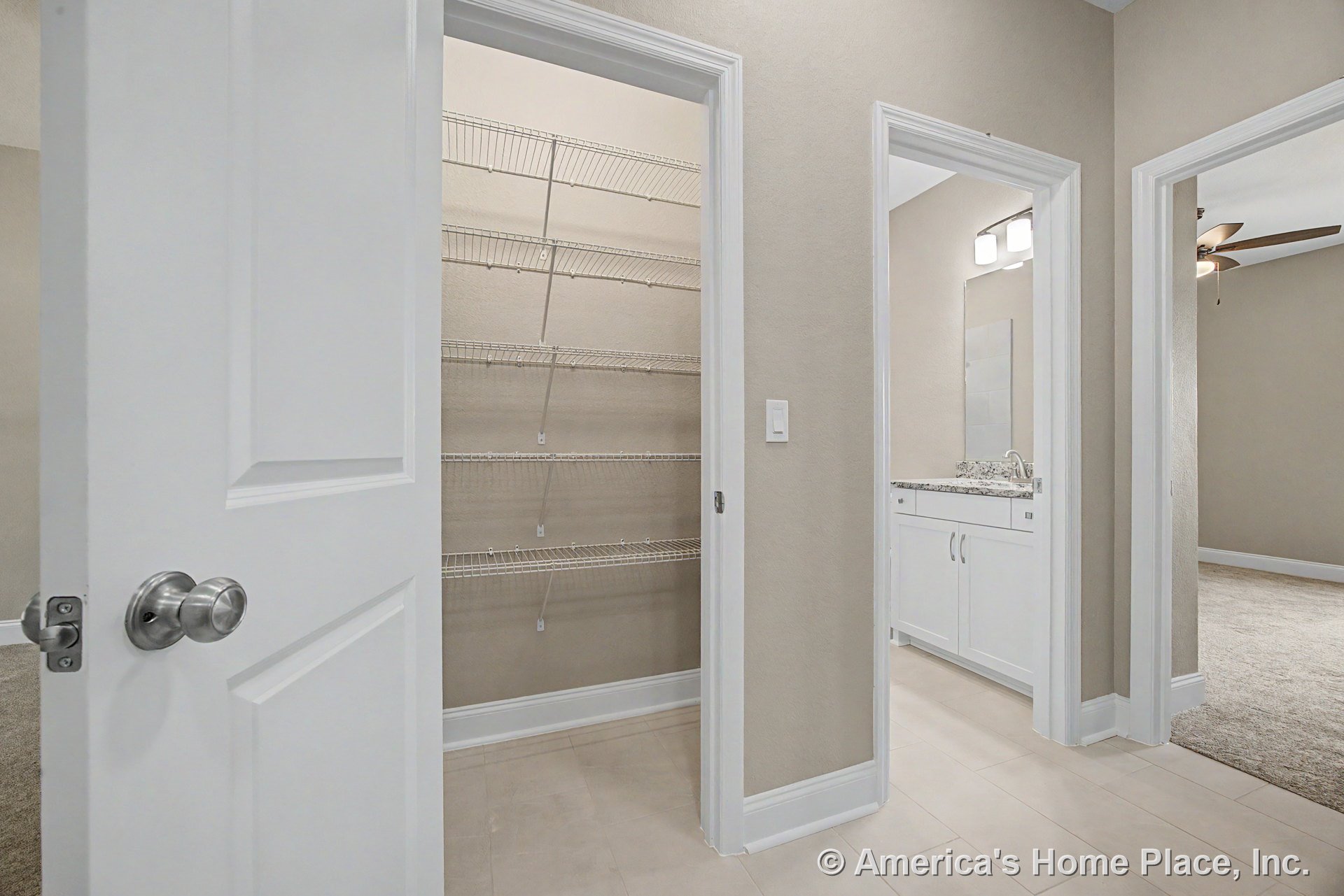 Hallway with tile flooring leads to a pantry with wire shelving, a bathroom featuring granite countertop and white cabinets, and a carpeted bedroom; neutral wall paint and built-in