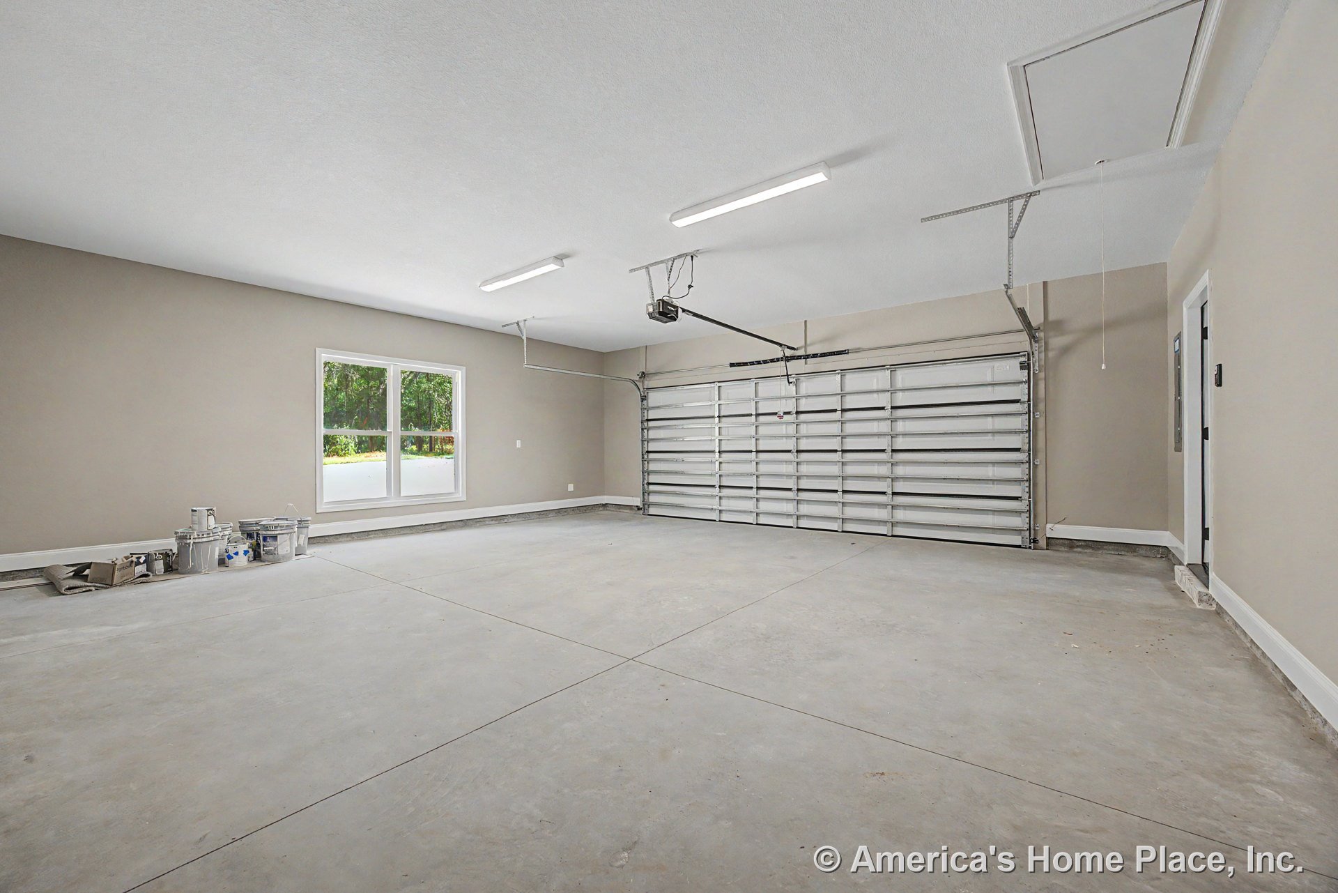 Double bay garage with concrete slab floor, large window, overhead fluorescent lighting, pull-down attic access, painted drywall walls with baseboard trim, and interior door.