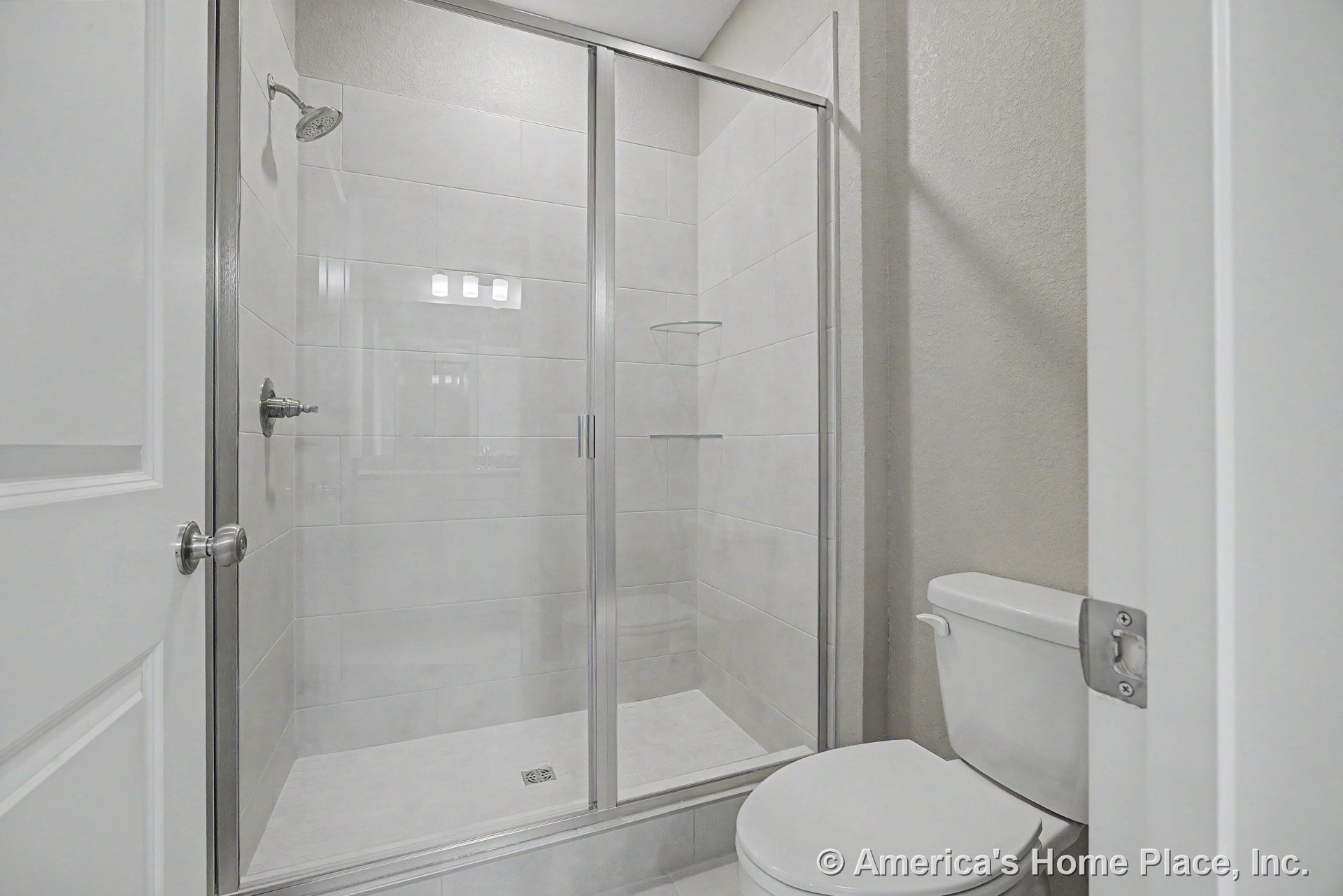 Walk-in shower with frameless glass enclosure, large light gray tile walls, built-in shower shelves, chrome showerhead, white toilet, and recessed ceiling.