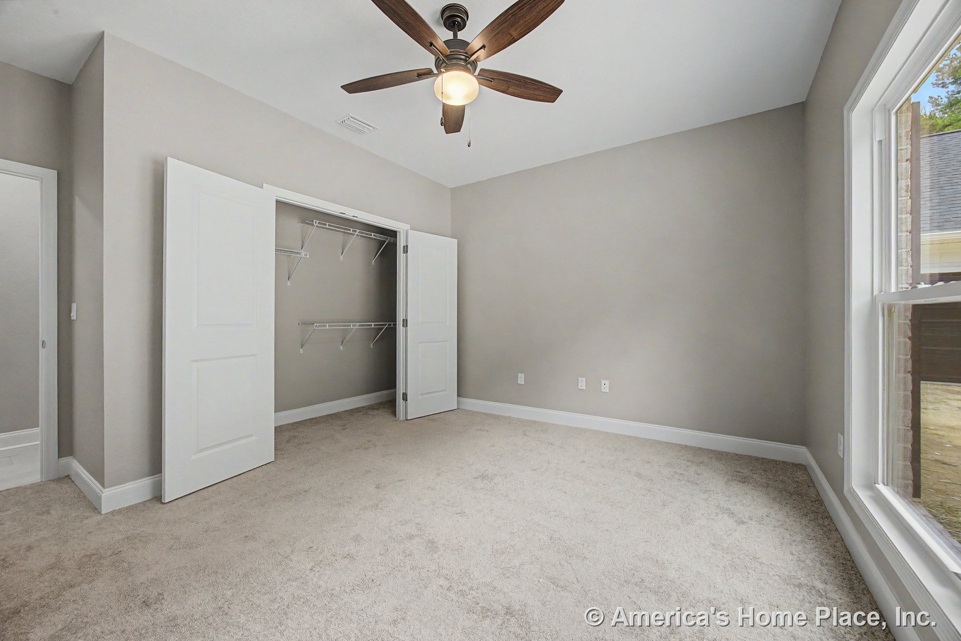 Bedroom with open closet featuring double doors and wire shelving, neutral carpet flooring, large window with white trim, ceiling fan with light fixture, light gray painted walls