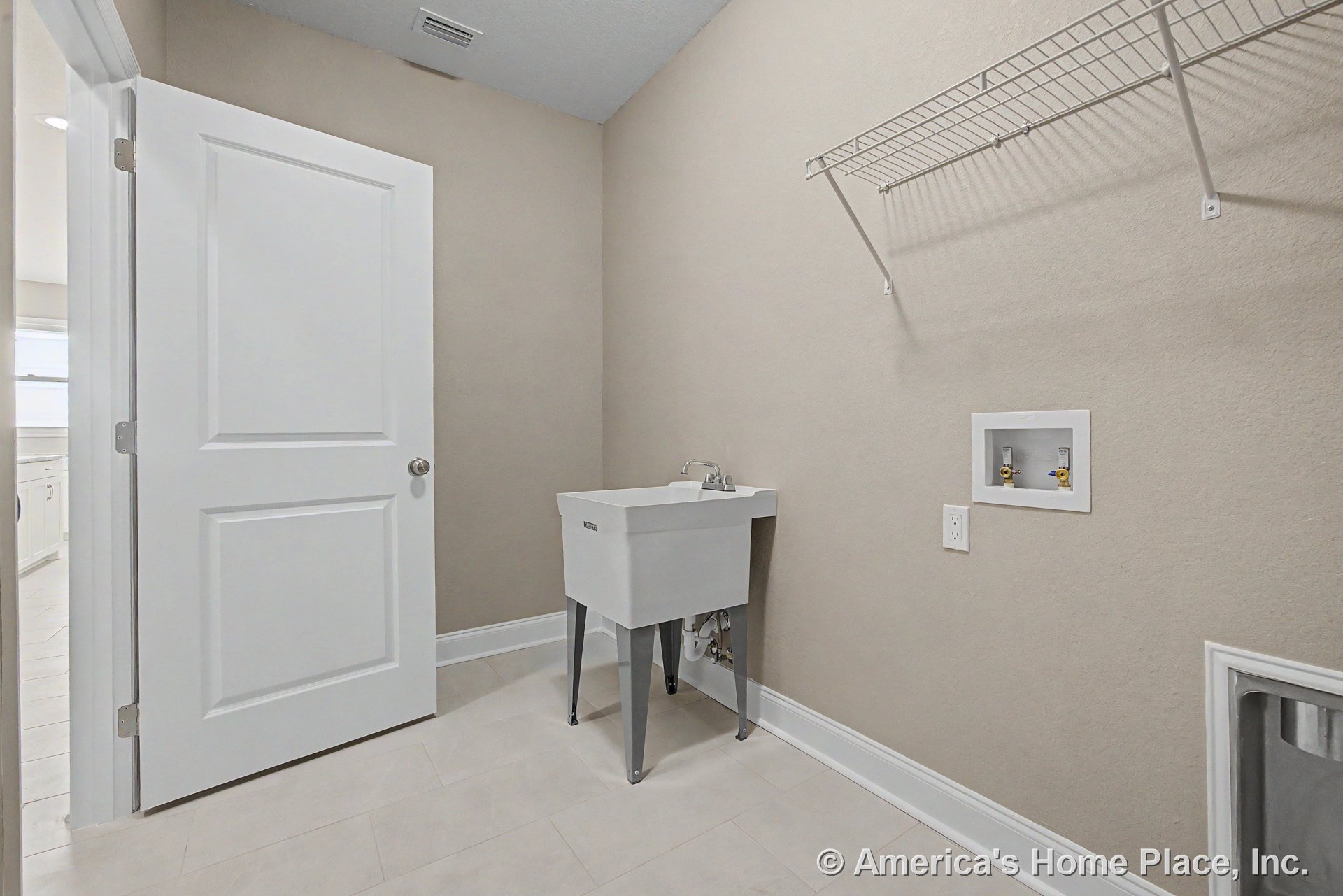 Utility sink with chrome faucet, washer and dryer hookups, white wire shelving for storage, beige painted walls, tile flooring, white paneled door, and baseboard trim.