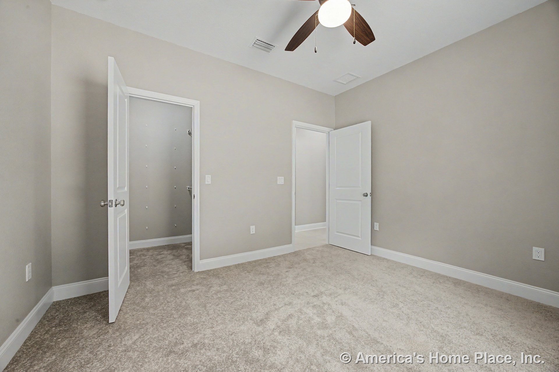 Beige carpet flooring, light gray walls with white trim and baseboards, ceiling fan with built-in light fixture, double panel doors, and walk-in closet in a neutral-toned bedroom.