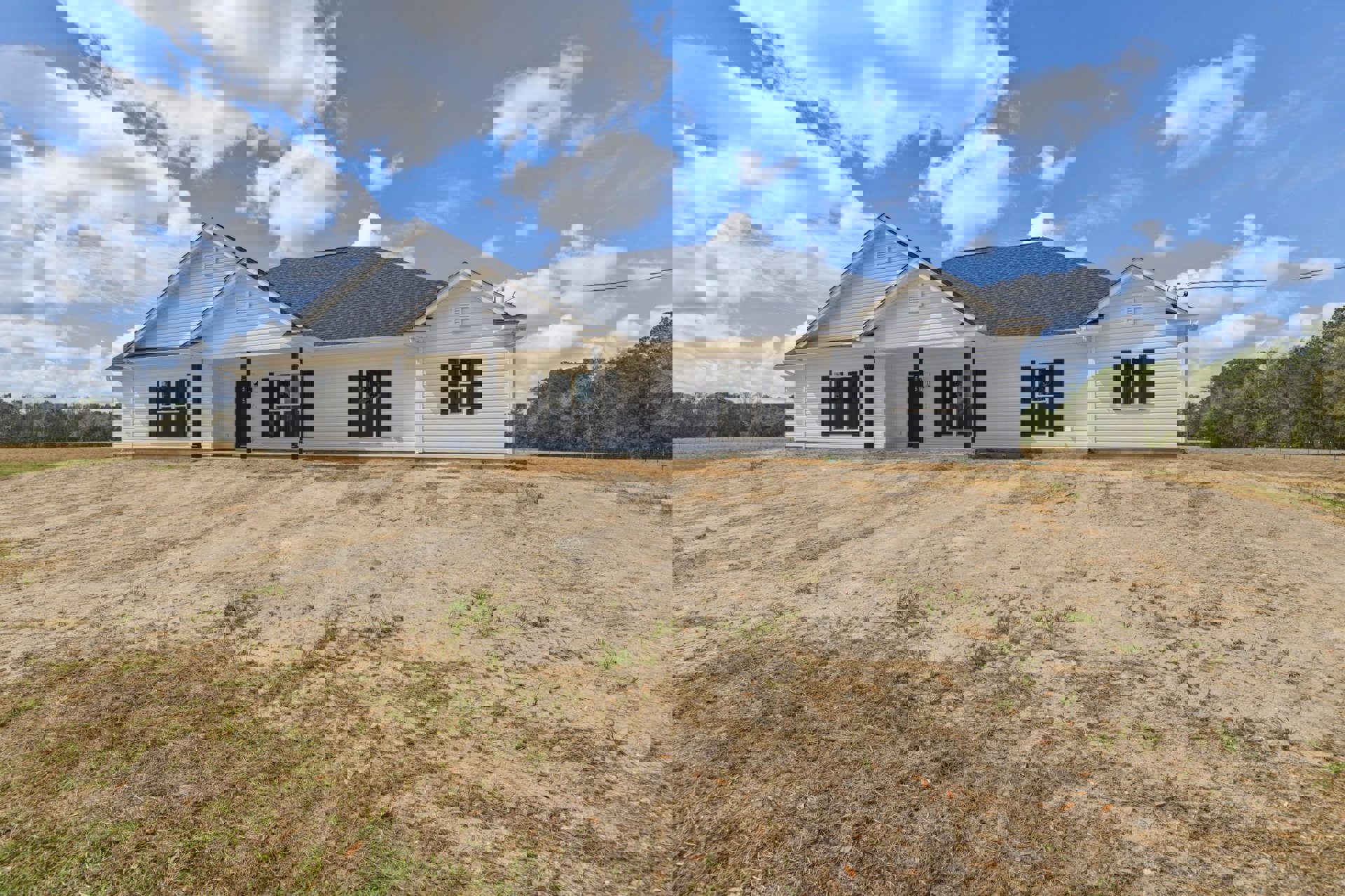 Two-story house with black roof, white-framed windows, and dirt field bordered by grass under blue sky with scattered clouds