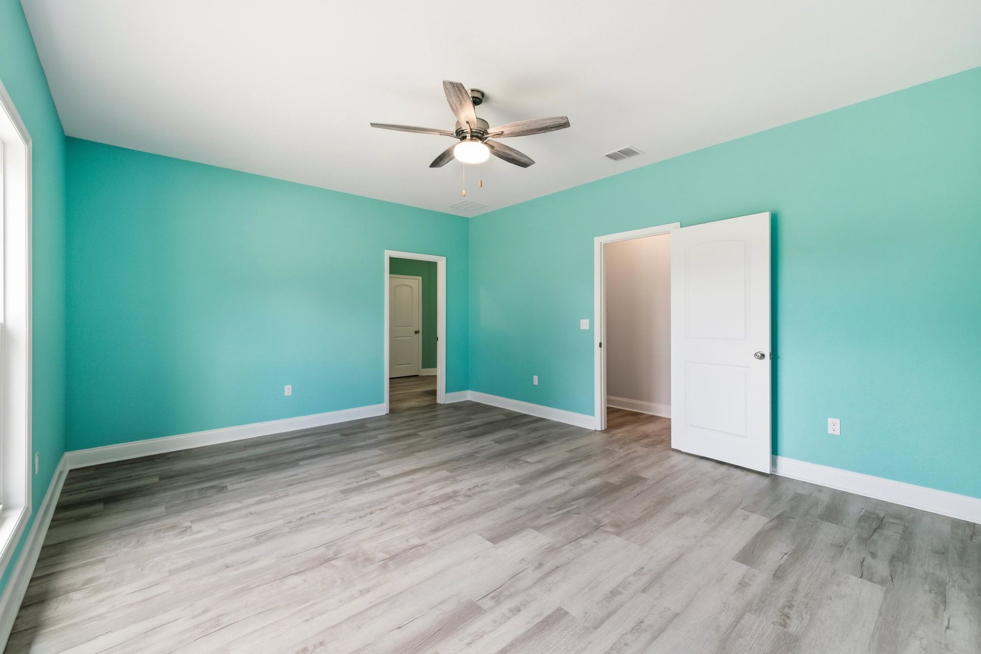 Blue-painted walls, wood laminate flooring, white door with silver knob, ceiling fan with light fixture, simple trim and plaster finishes