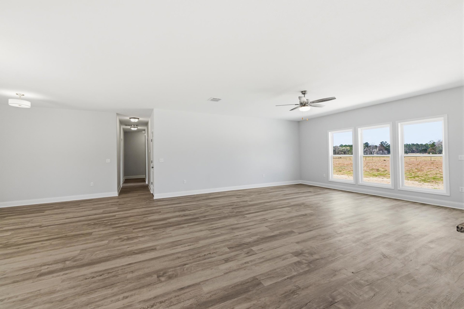 Spacious room with wood flooring, white walls, ceiling fan with light, large windows overlooking grassy field, and door with wall-mounted light fixture