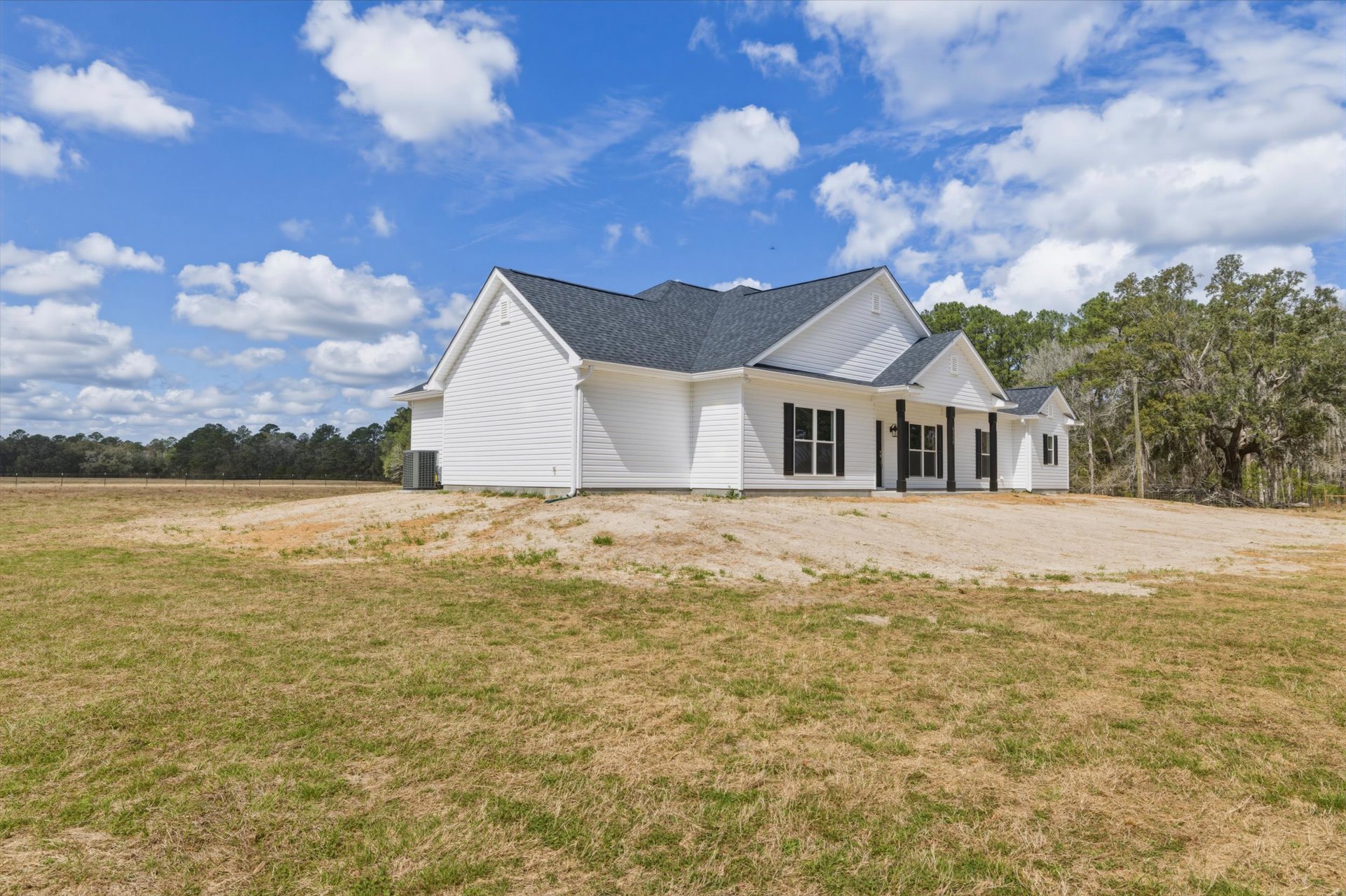 White two-story house with black shuttered windows, gray roof, and surrounded by a grassy field under a blue sky with scattered clouds, bordered by trees.