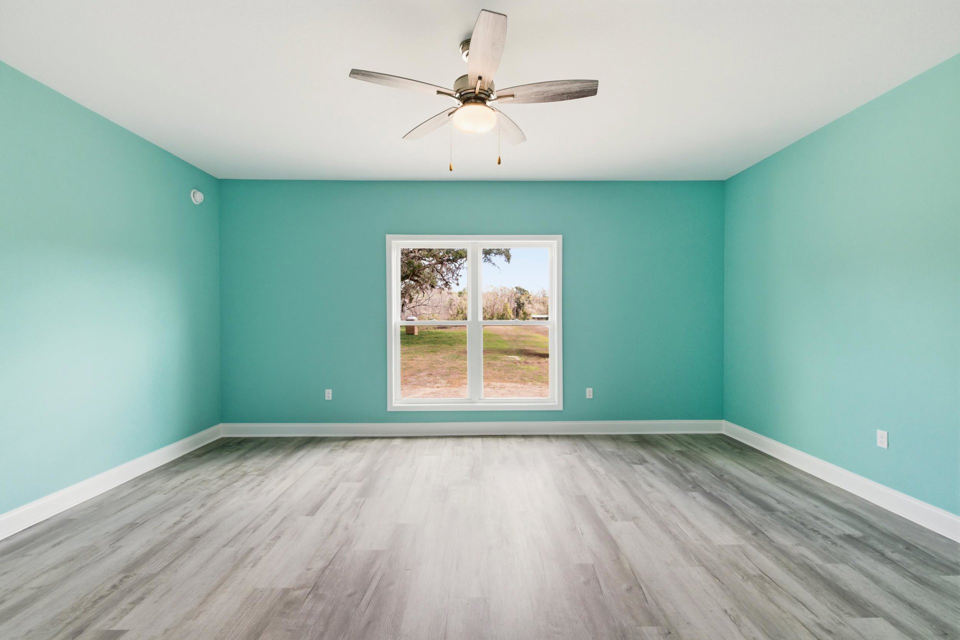 Blue accent wall and white wood flooring in a room featuring a ceiling fan with light, large window overlooking a grassy field.
