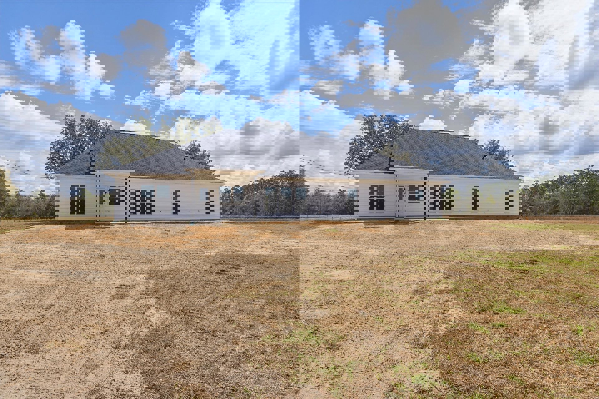 White house with black roof set behind a large patch of brown dirt, surrounded by grassy areas and trees under a blue sky.