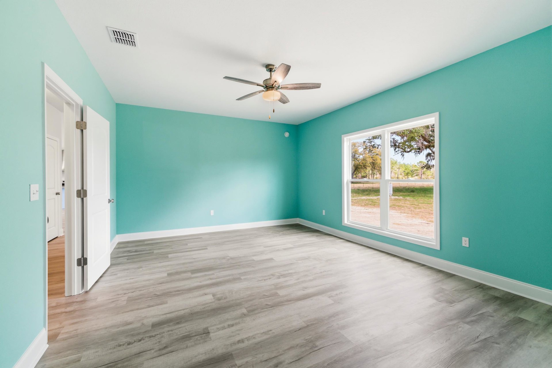 Ceiling fan with light fixture, large window overlooking grassy field, wood laminate flooring, white plaster walls, wall vent
