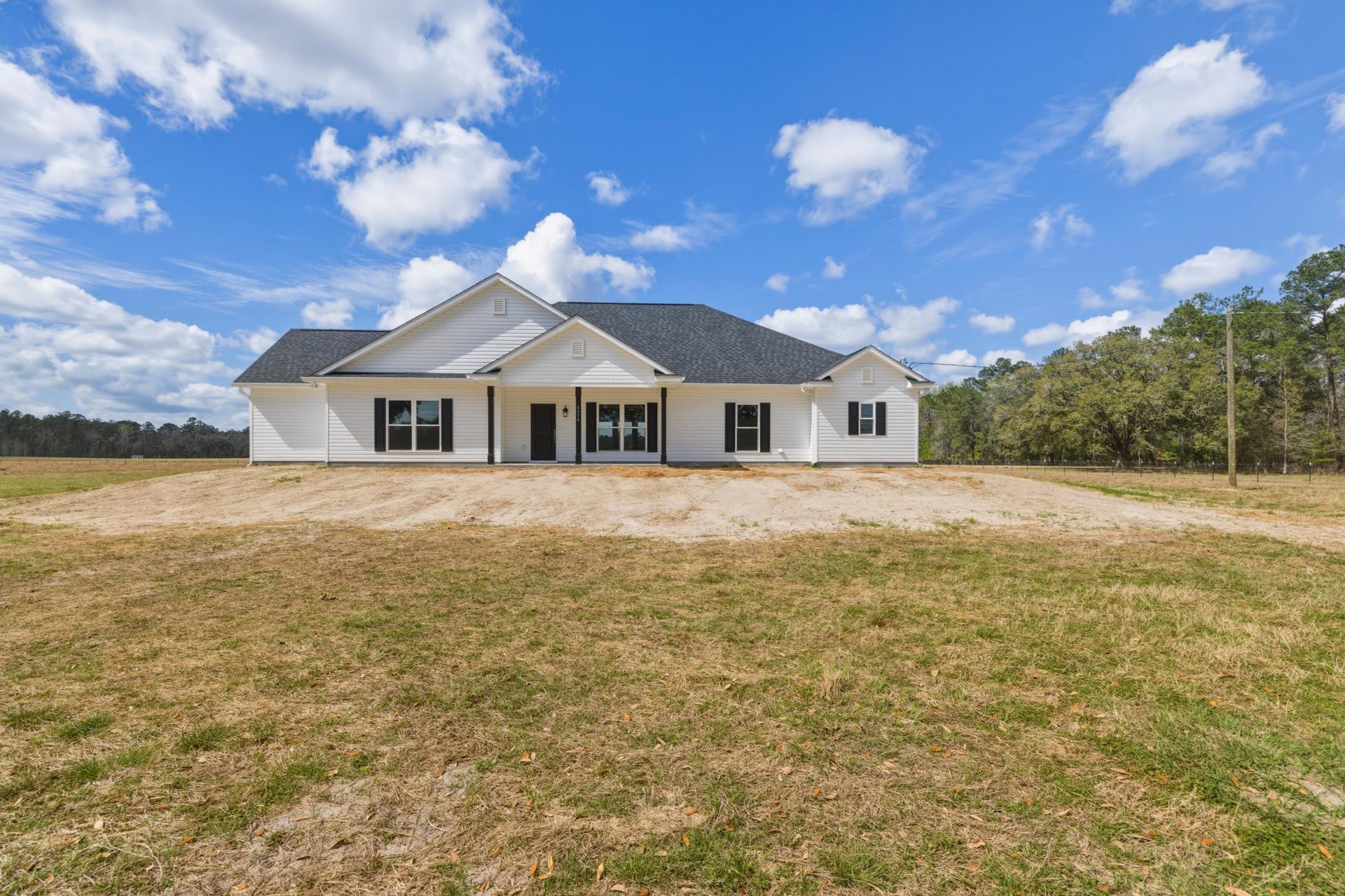 White house with black shutters, large grassy yard, dirt driveway, blue sky with scattered clouds, mature trees surrounding property