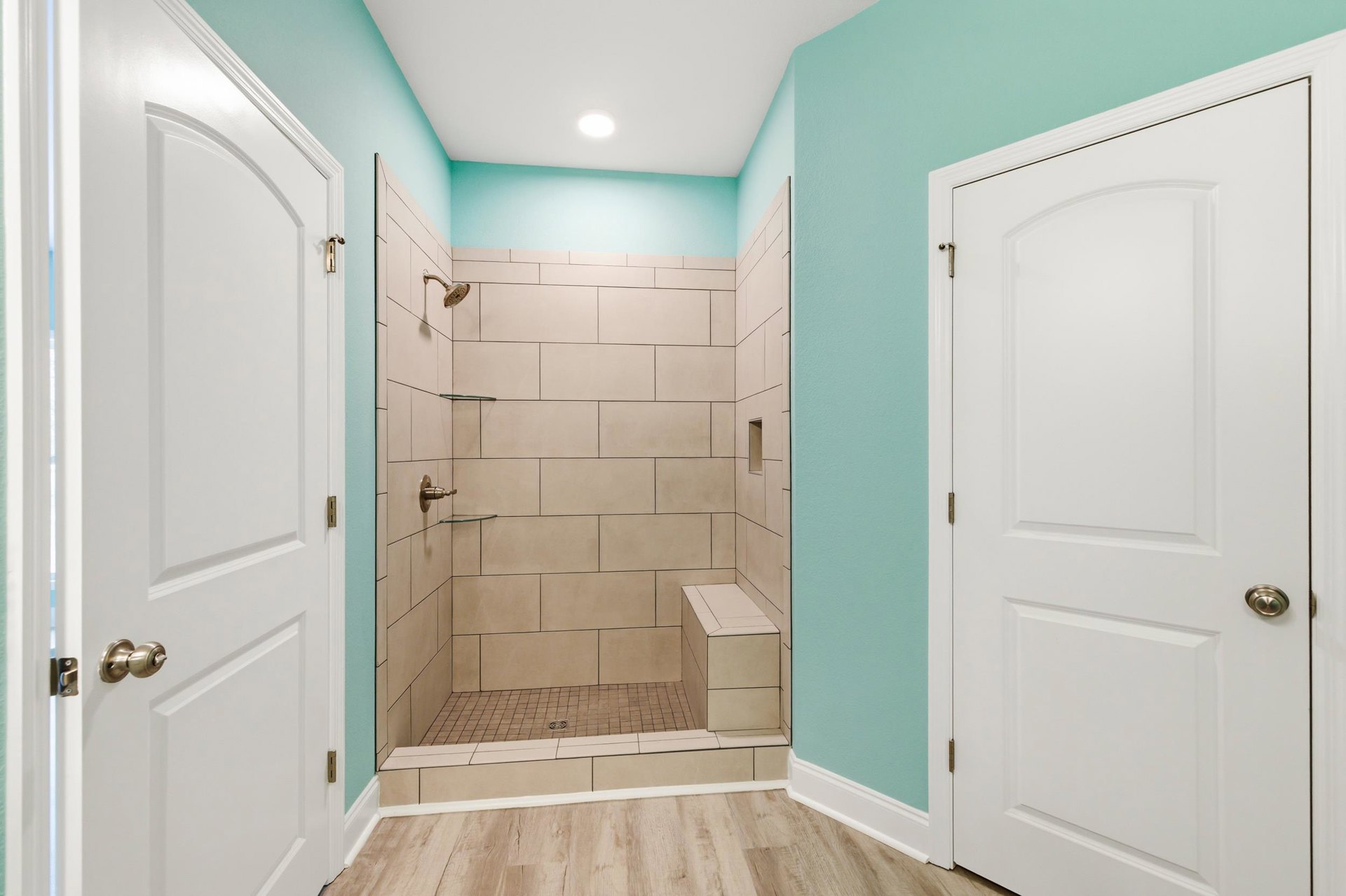 Bathroom with wood flooring, white doors featuring brass handles, glass-enclosed shower with built-in bench and chrome showerhead, neutral wall finishes.