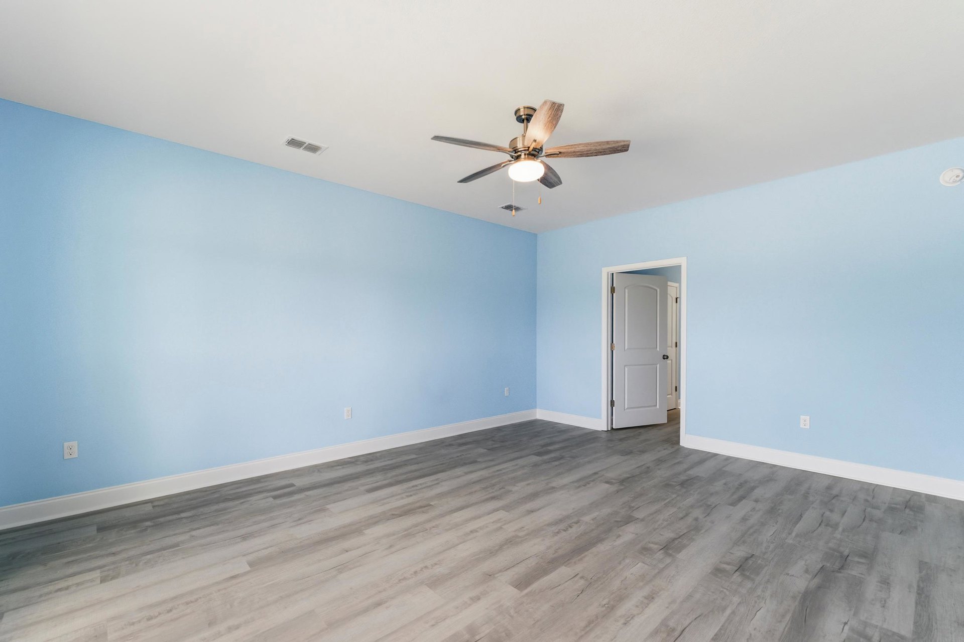 Ceiling fan with light fixture, blue accent wall, grey wood laminate flooring, white door with black handle, white baseboards and trim