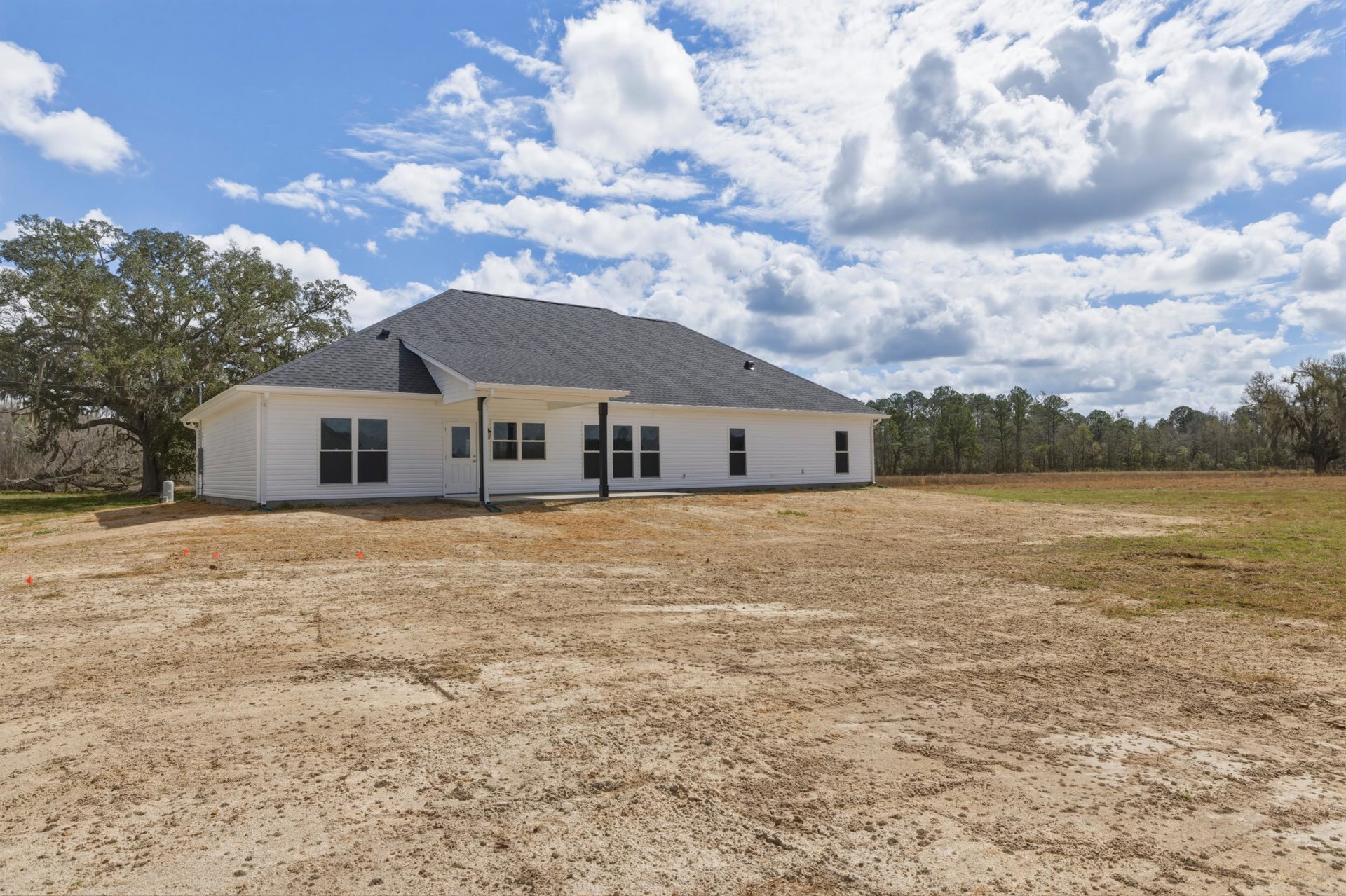 White house with black roof set on a dirt field, surrounded by trees under a blue sky with clouds
