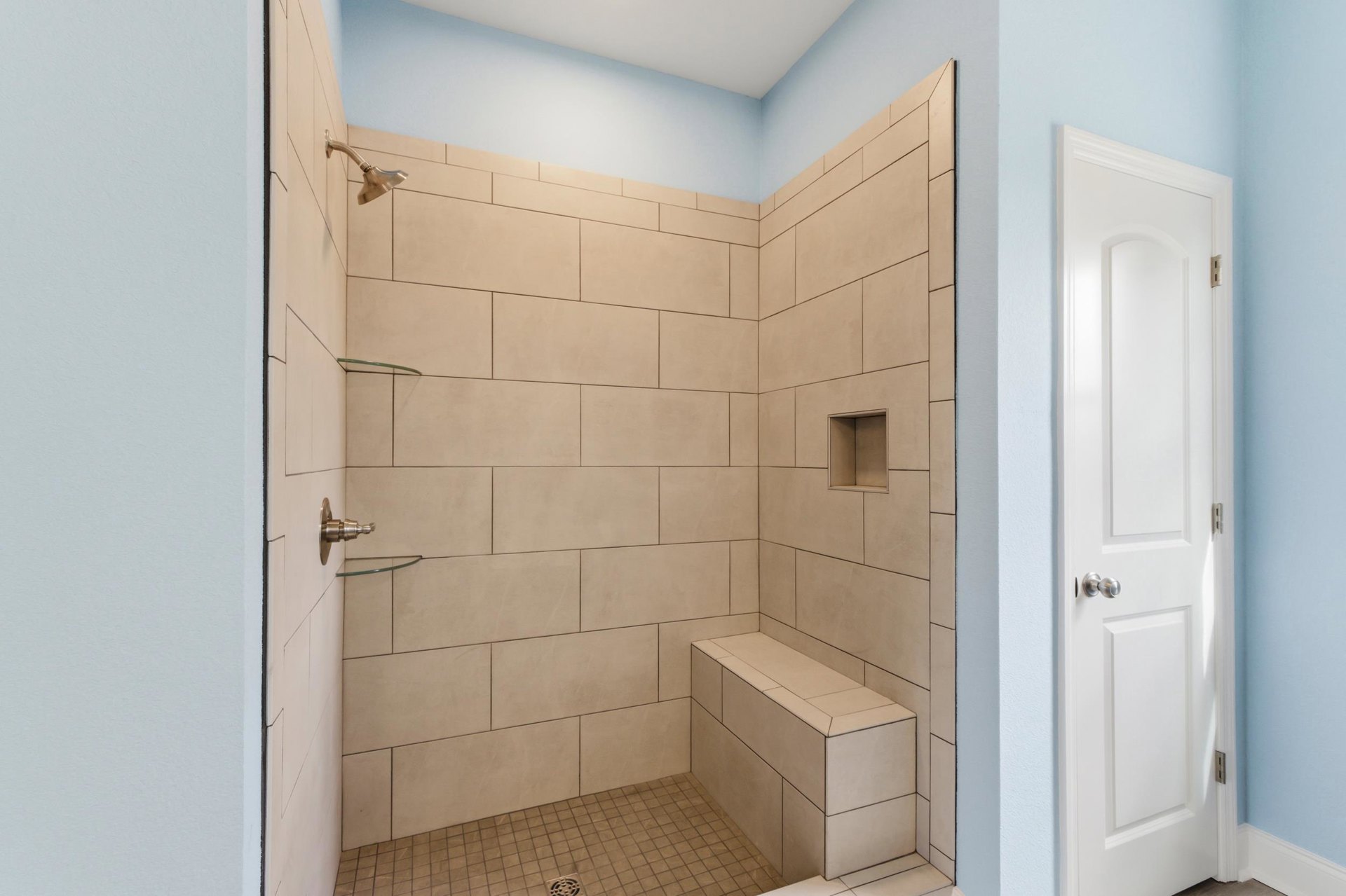 White tiled shower with built-in bench and seat, silver shower head, adjacent white door with silver knob, square white shelf.