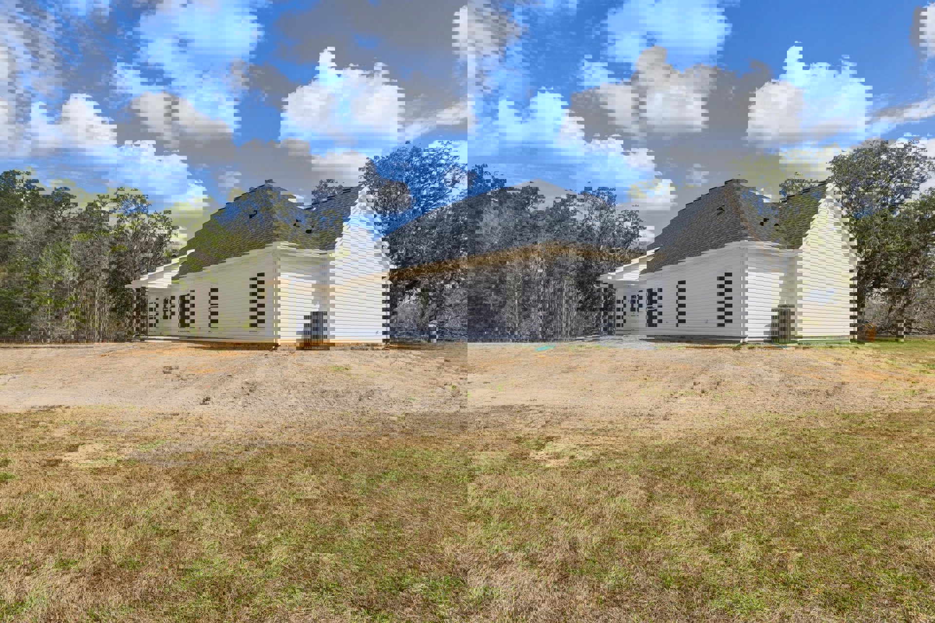 White farmhouse with black roof, surrounded by grassy field, large tree in background, grey metal tower nearby, blue sky with scattered clouds