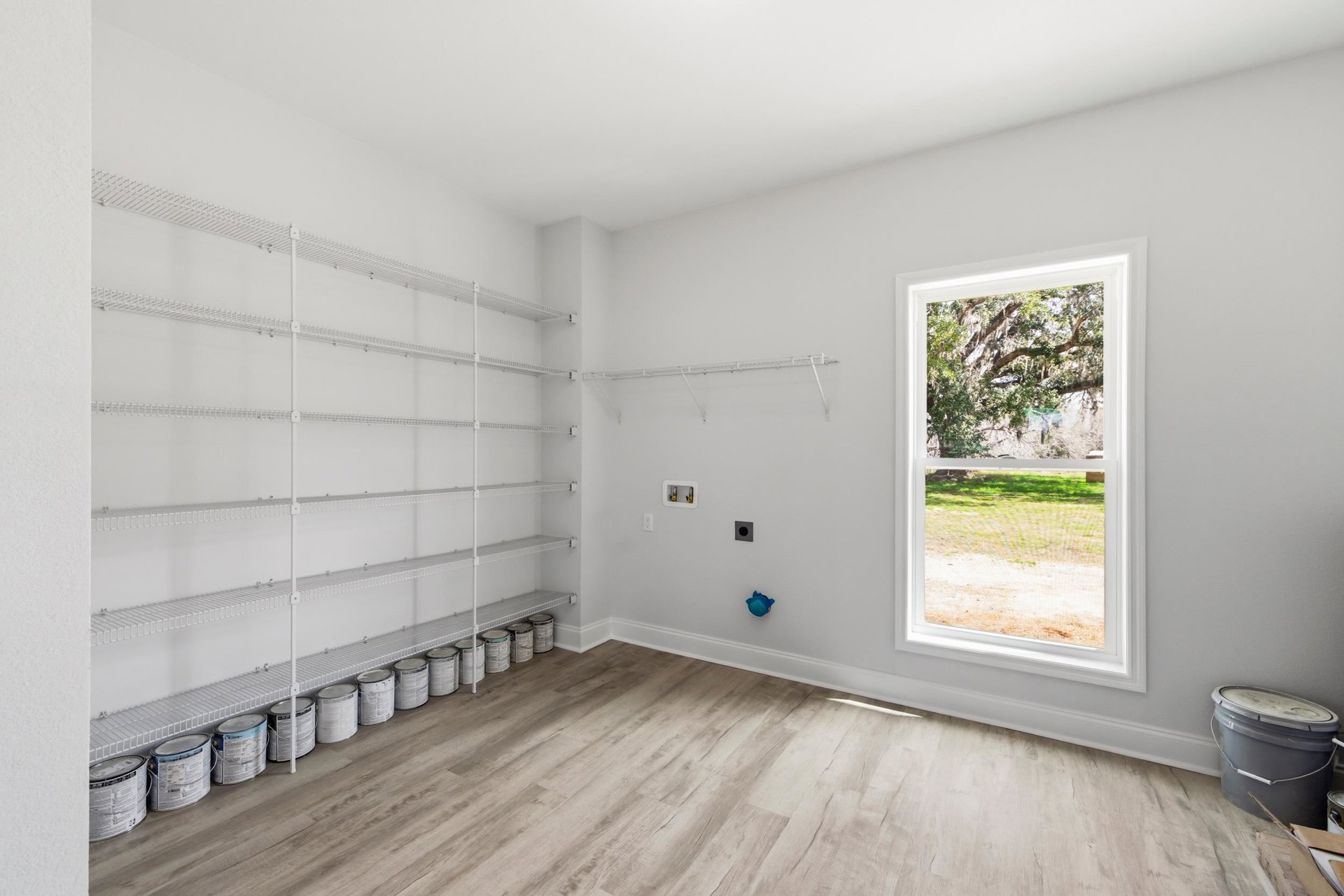 Built-in shelving and storage buckets along a plaster wall, wood flooring, window overlooking yard and trees, paint can and blue bucket in corner