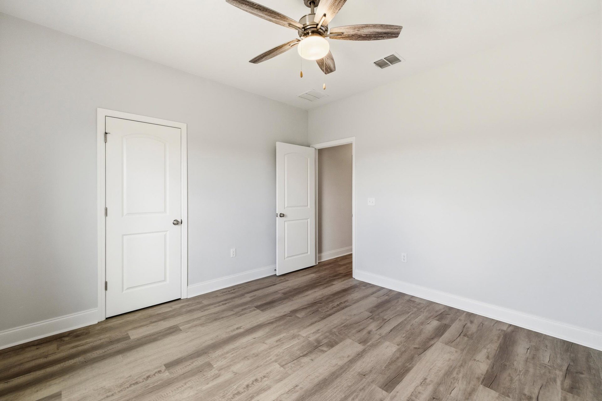 Ceiling fan with light fixture, two white doors with silver knobs, wood flooring, white walls, and ceiling vent