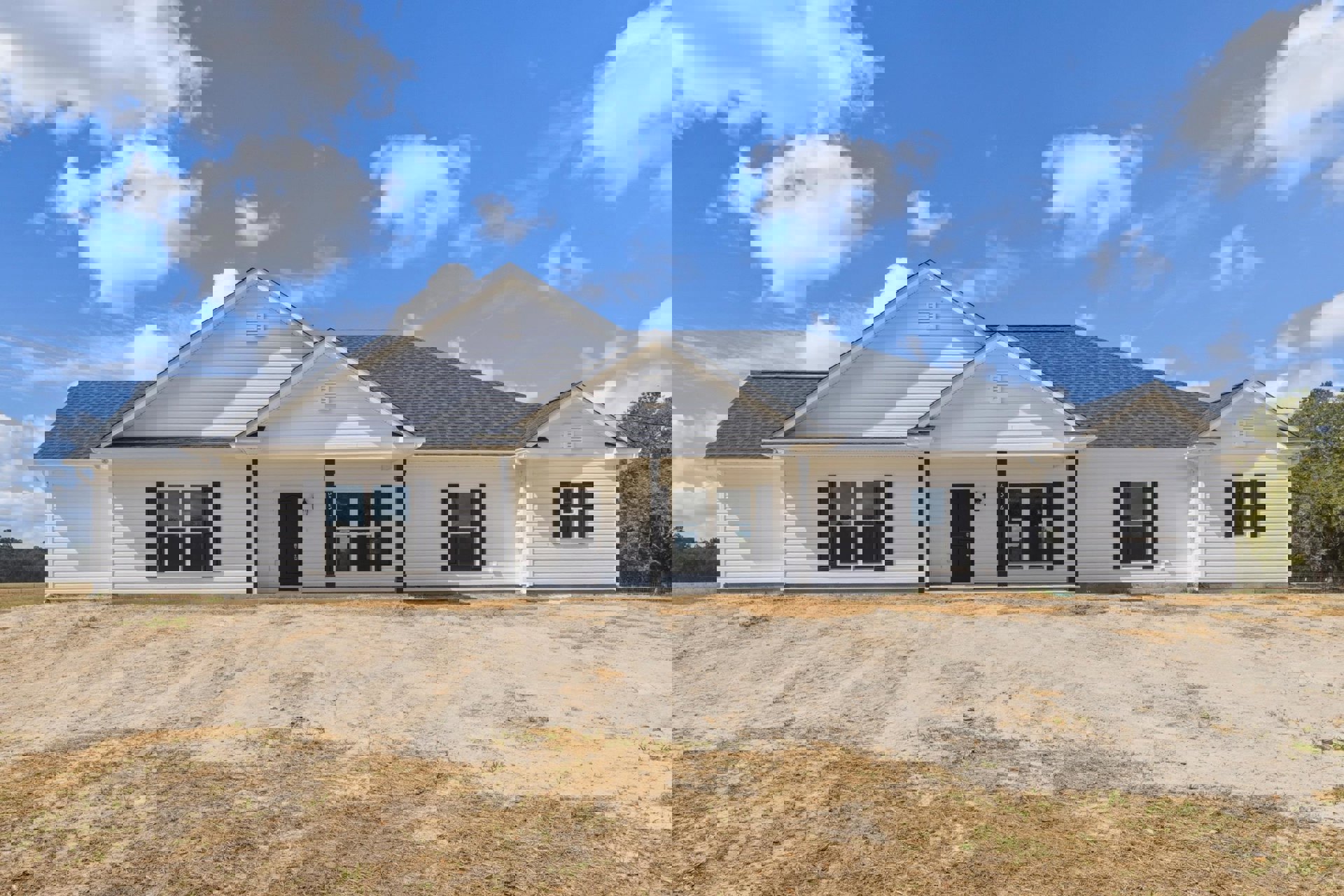 White house with black shutters and white-framed windows, black front door with silver handle, dirt road and grassy field in foreground, blue sky with clouds above