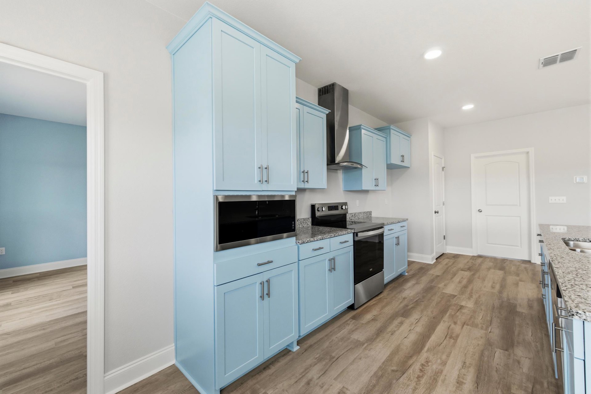 Blue kitchen cabinets with silver hardware, wood plank flooring, black and silver microwave above matching dishwasher, white door with silver handle, blue walls with white trim.