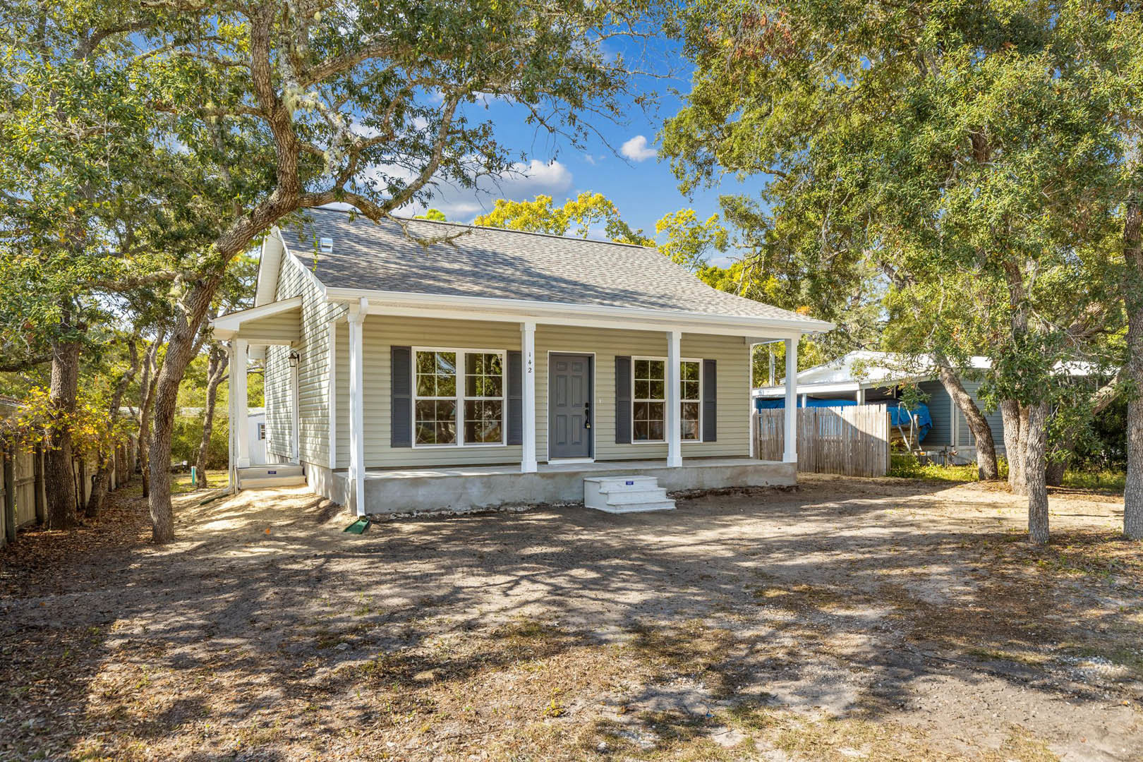 Two-story house surrounded by mature trees, grey front door with lock, covered porch, multi-pane windows, dirt yard with white concrete step, white rectangular utility box with