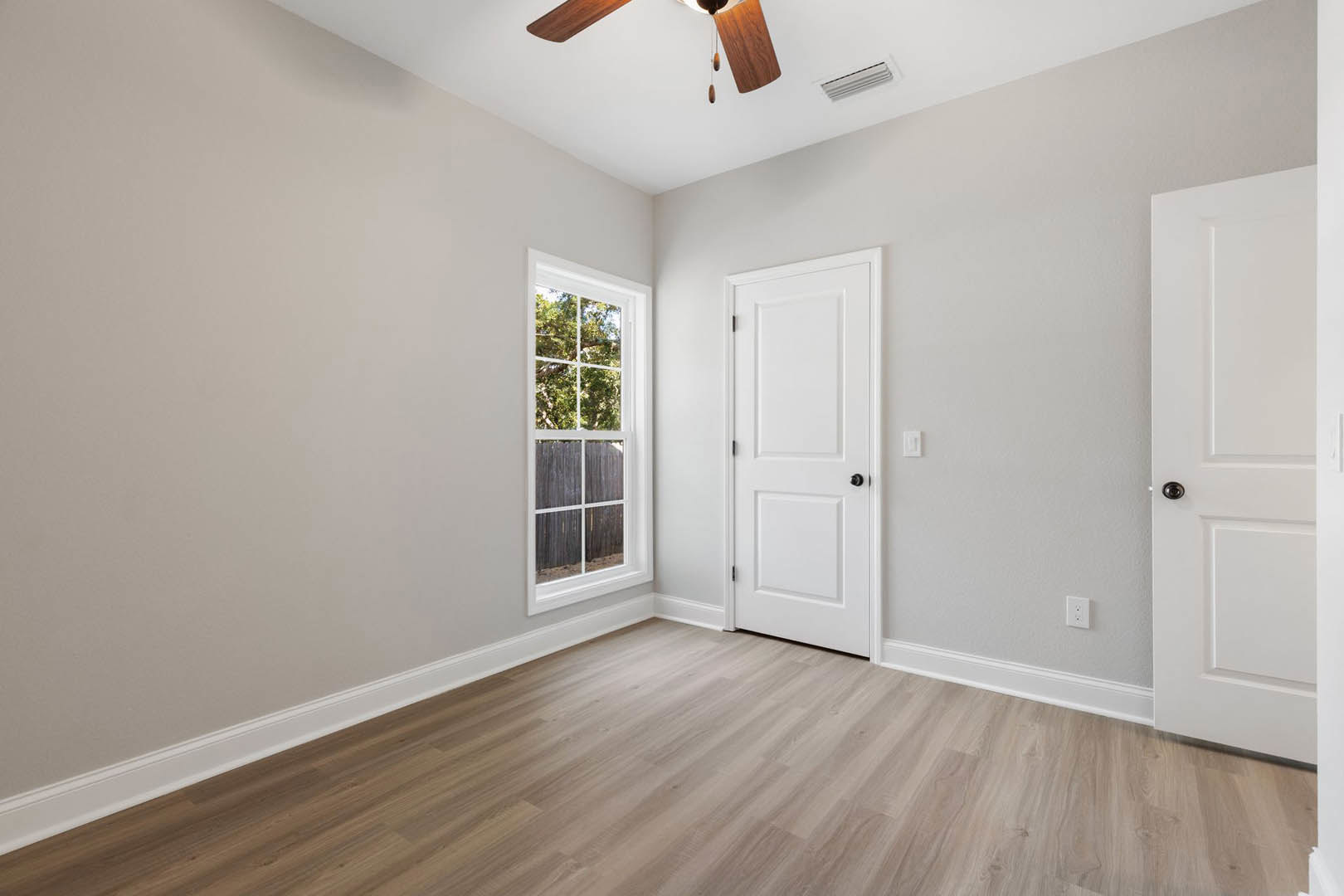 Ceiling fan mounted above hardwood floor, white door with black knob, window overlooking trees, neutral walls