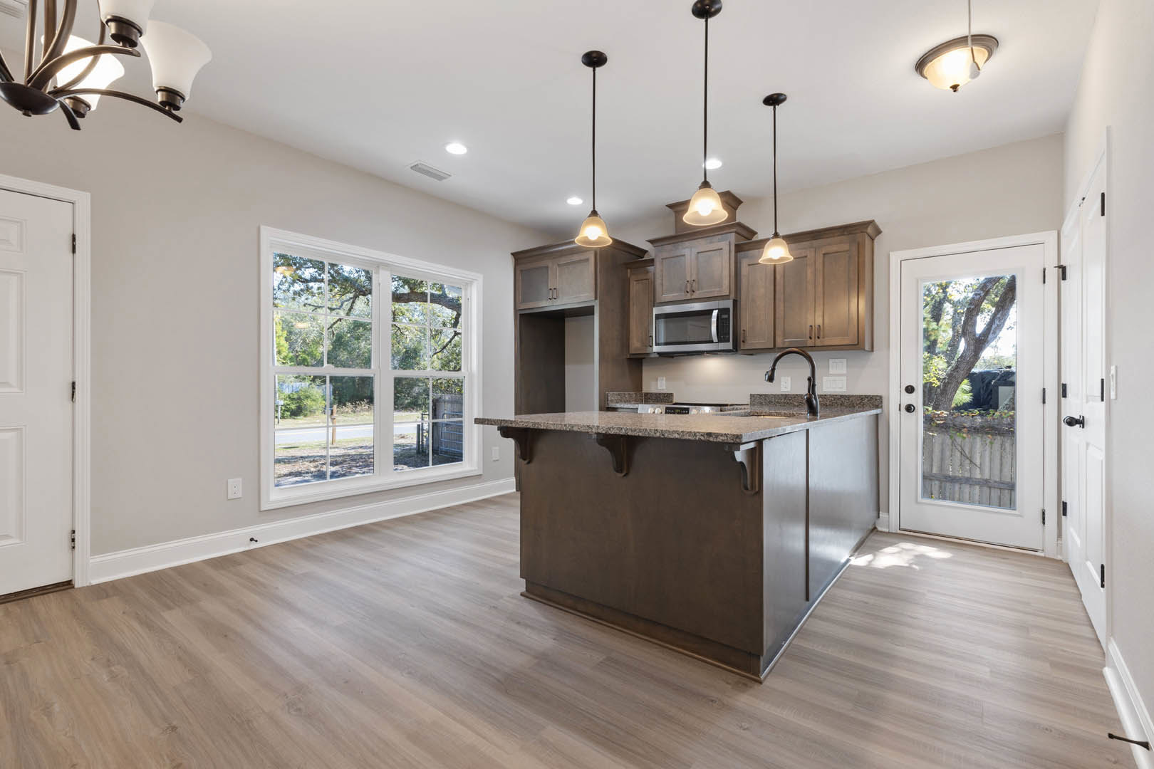 Kitchen with wood flooring, marble-topped island, built-in microwave, bar seating, white cabinetry, recessed ceiling lights, large window and glass door offering tree views.