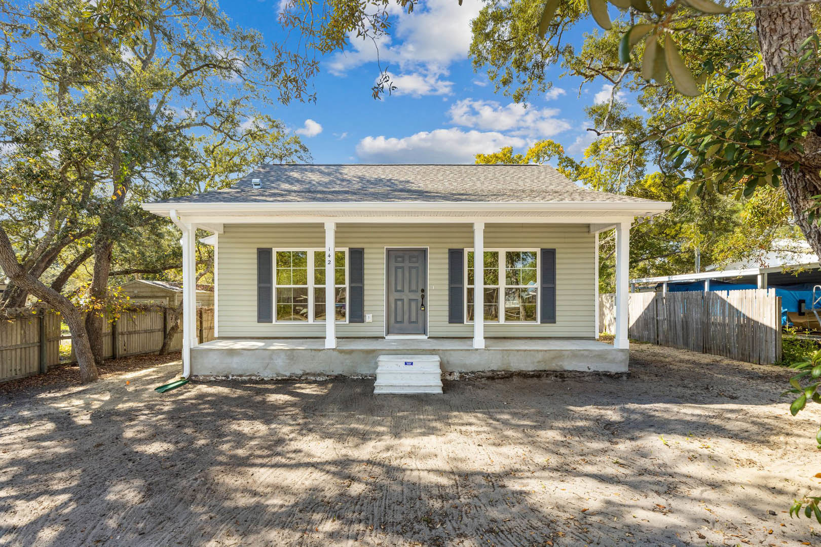 Two-story house with covered porch, white steps, grey door, wooden fence, blue roof accents, and dirt ground surrounded by trees and cloudy sky