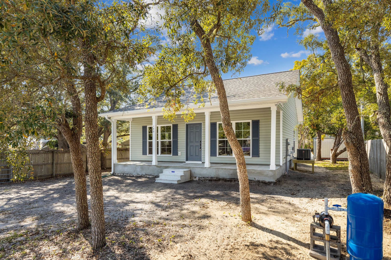 Modern home surrounded by mature trees, white framed windows, glass-paneled white door, blue barrel on bare dirt, stack of books on concrete porch, close-up of outdoor water pump