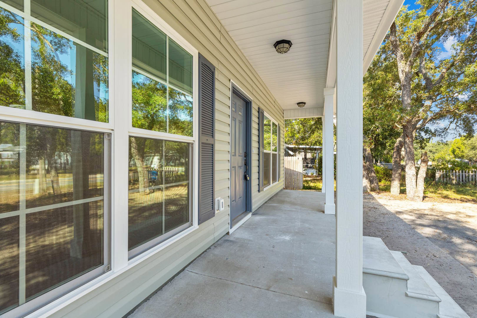 Covered front porch with wood ceiling, hanging light fixture, white door, large windows reflecting trees, and landscaping with mature tree.