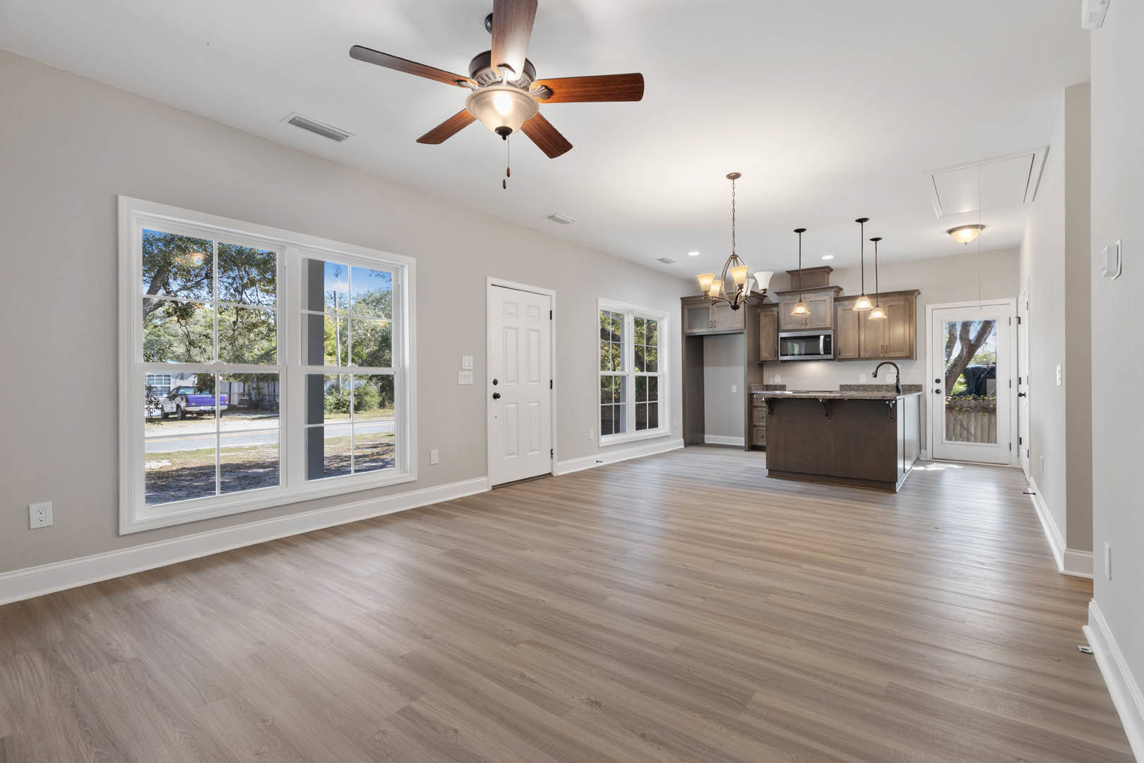 Open living room with wood laminate flooring, ceiling fan with light fixture, white door with black knobs, adjacent kitchen featuring stainless steel microwave, window overlooking