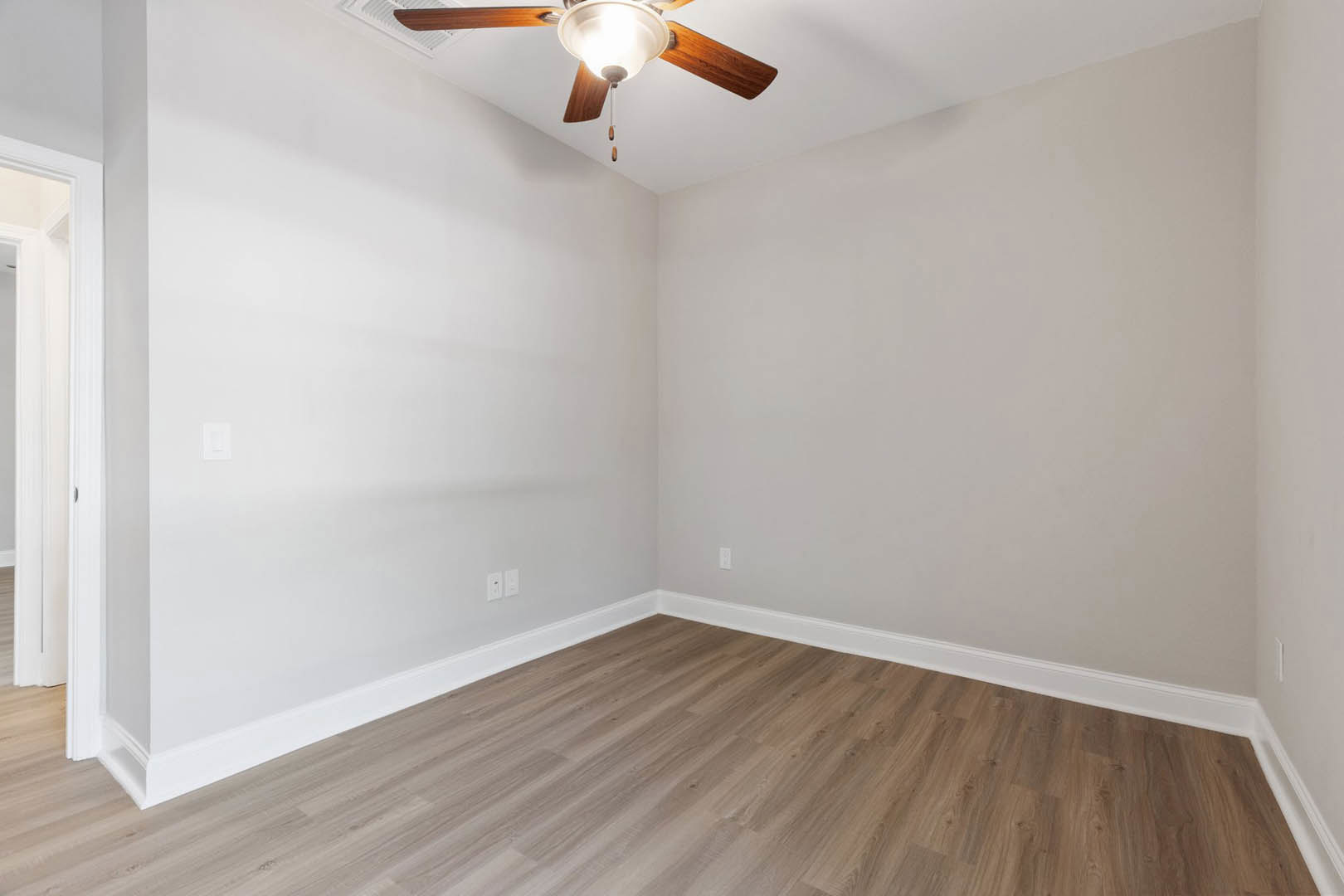 Ceiling fan with light fixture mounted on white plaster ceiling above hardwood floor with white baseboards, white wall outlet and light switch visible on wall.