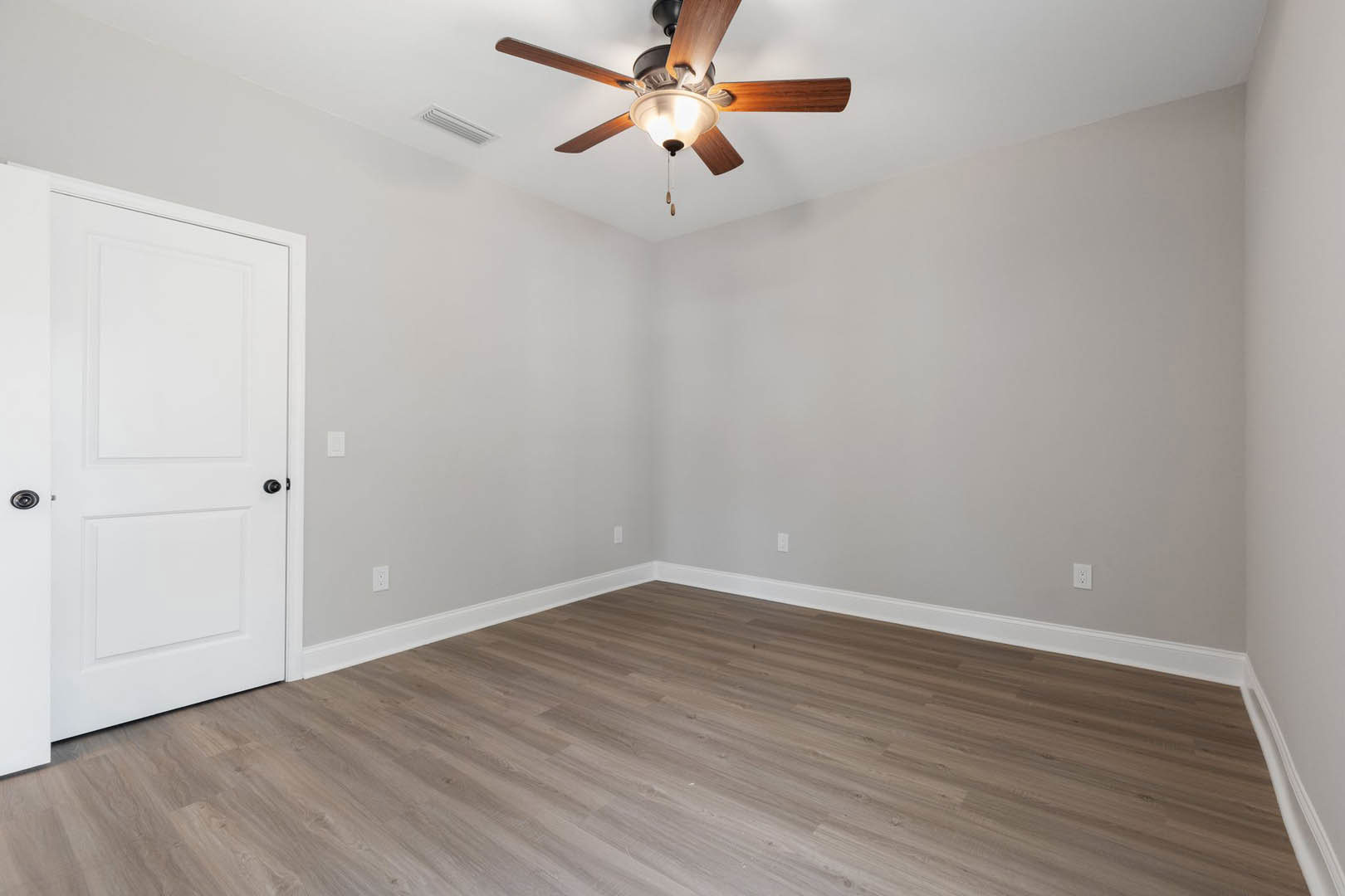 Ceiling fan with wooden blades and light fixture, wood flooring with white baseboards, white door with black hardware, smooth plaster walls