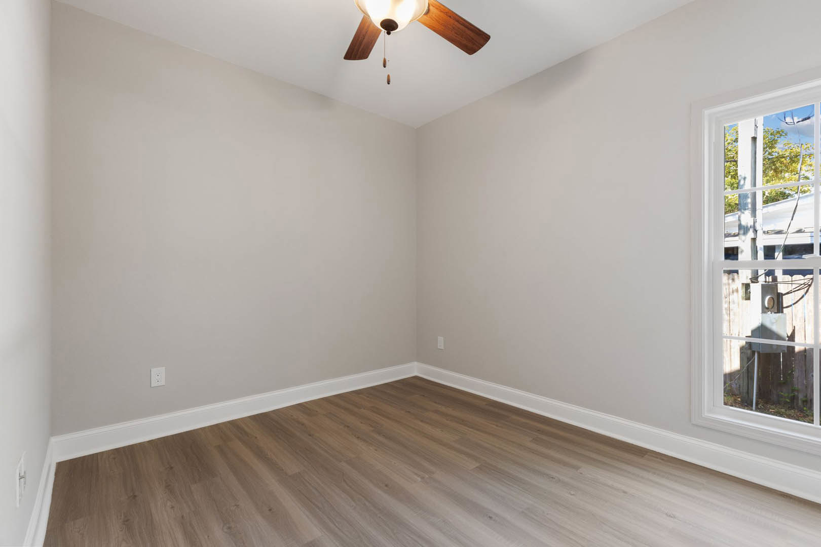 Ceiling fan with light fixture mounted on white plaster ceiling, wood flooring with white baseboards, large window with white frame, neutral painted walls