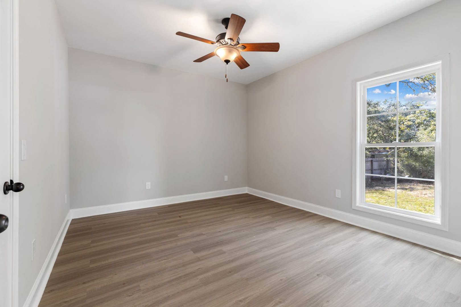 Wood flooring in a room with white walls, ceiling fan with light fixture, large window showing trees outside, close-up of brushed metal door knob