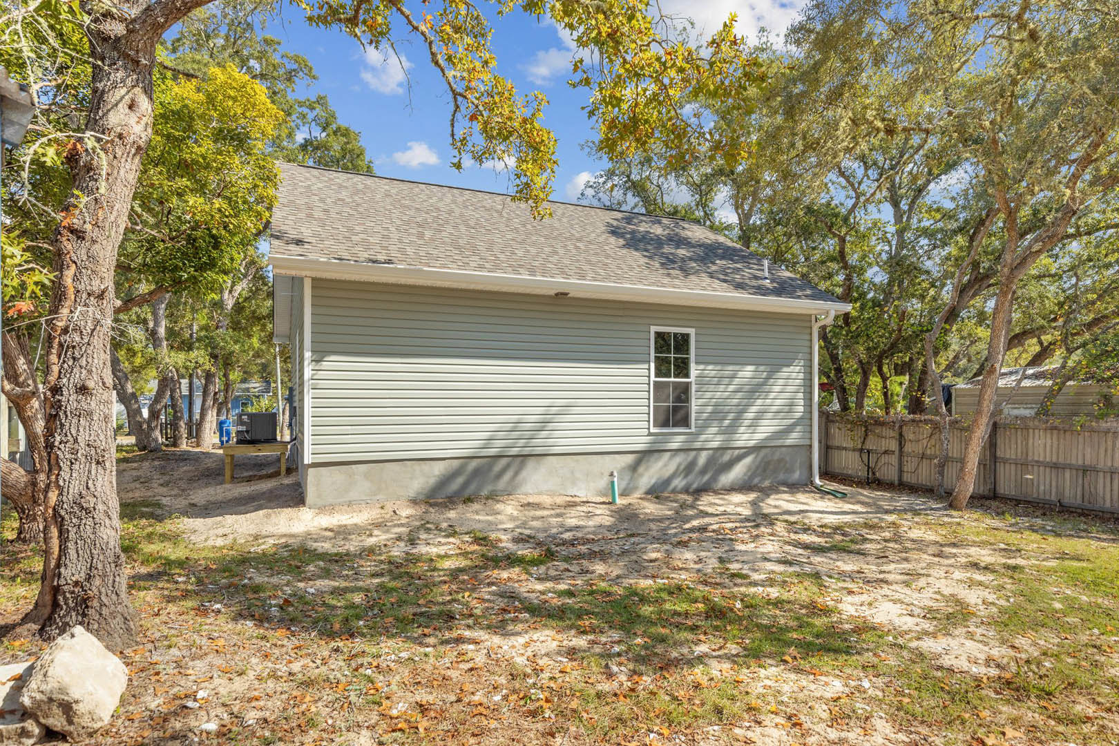 Two-story home with white-framed windows, light siding, grassy yard scattered with leaves, large rock, wooden table, and mature trees in the background