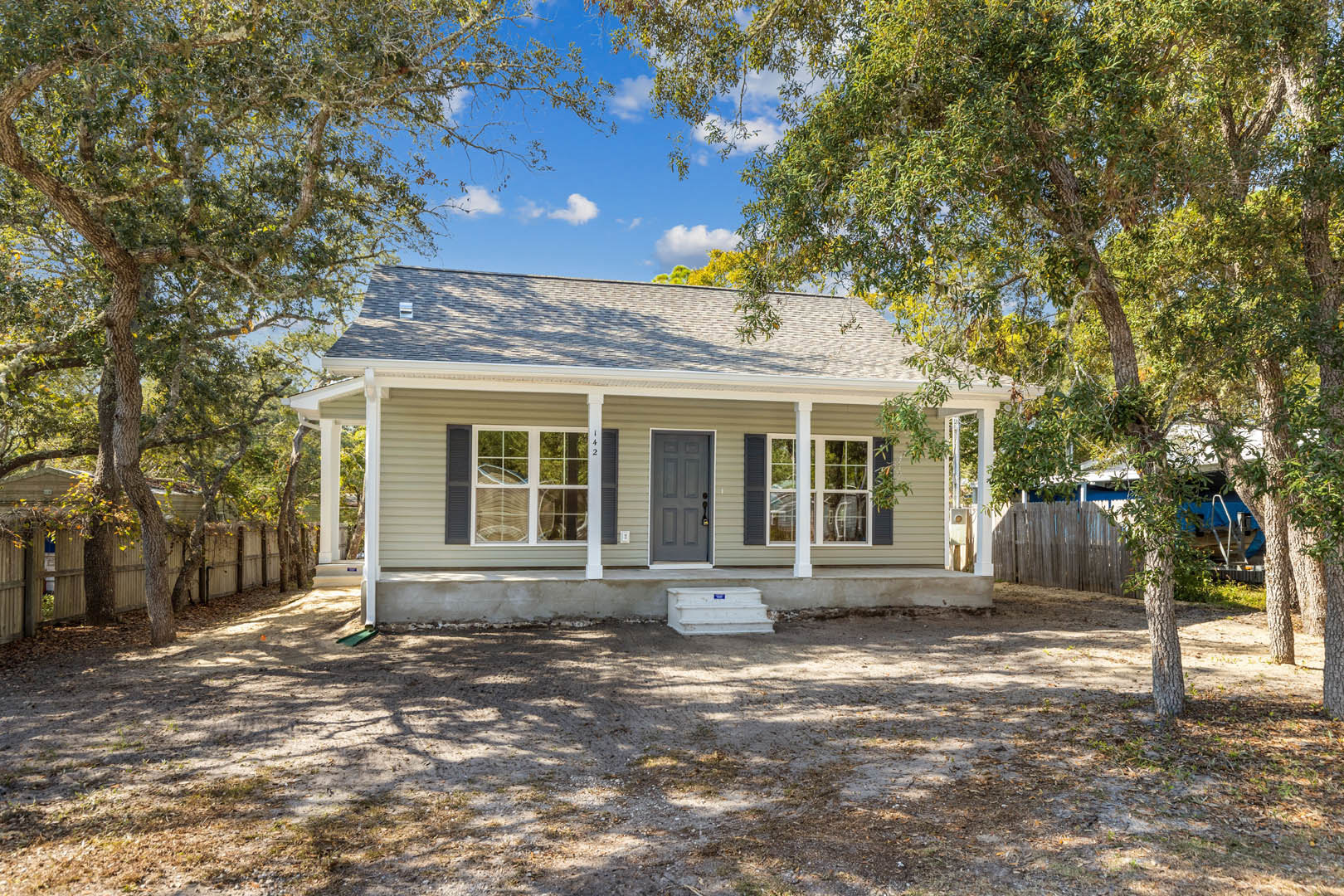 Grey front door with white pillars, white-framed window, dirt ground with white step, surrounded by mature trees under blue sky