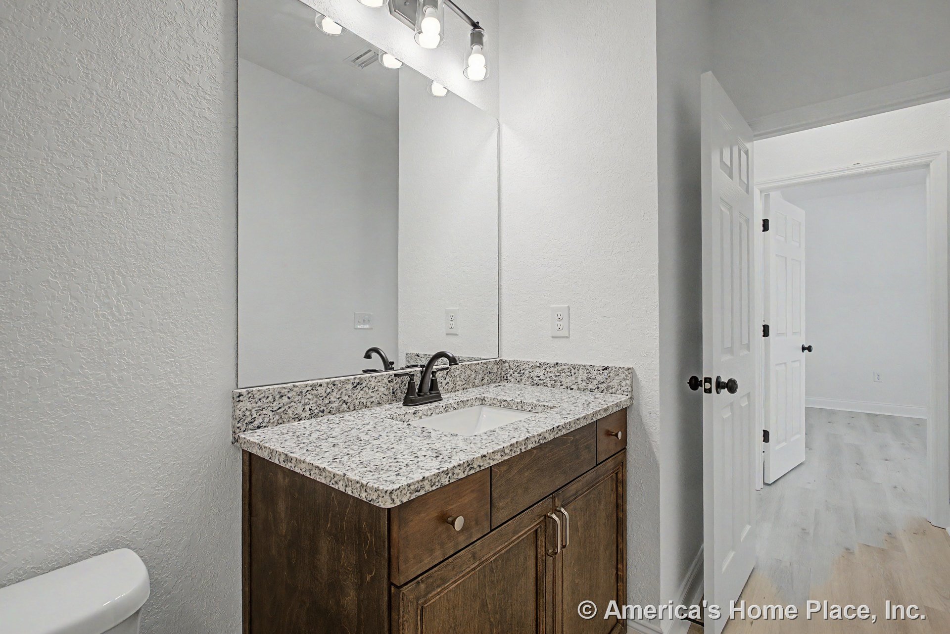 Granite countertop vanity with dark wood cabinetry, large wall mirror, modern light fixture, white textured walls, interior door with black hardware, undermount sink and chrome