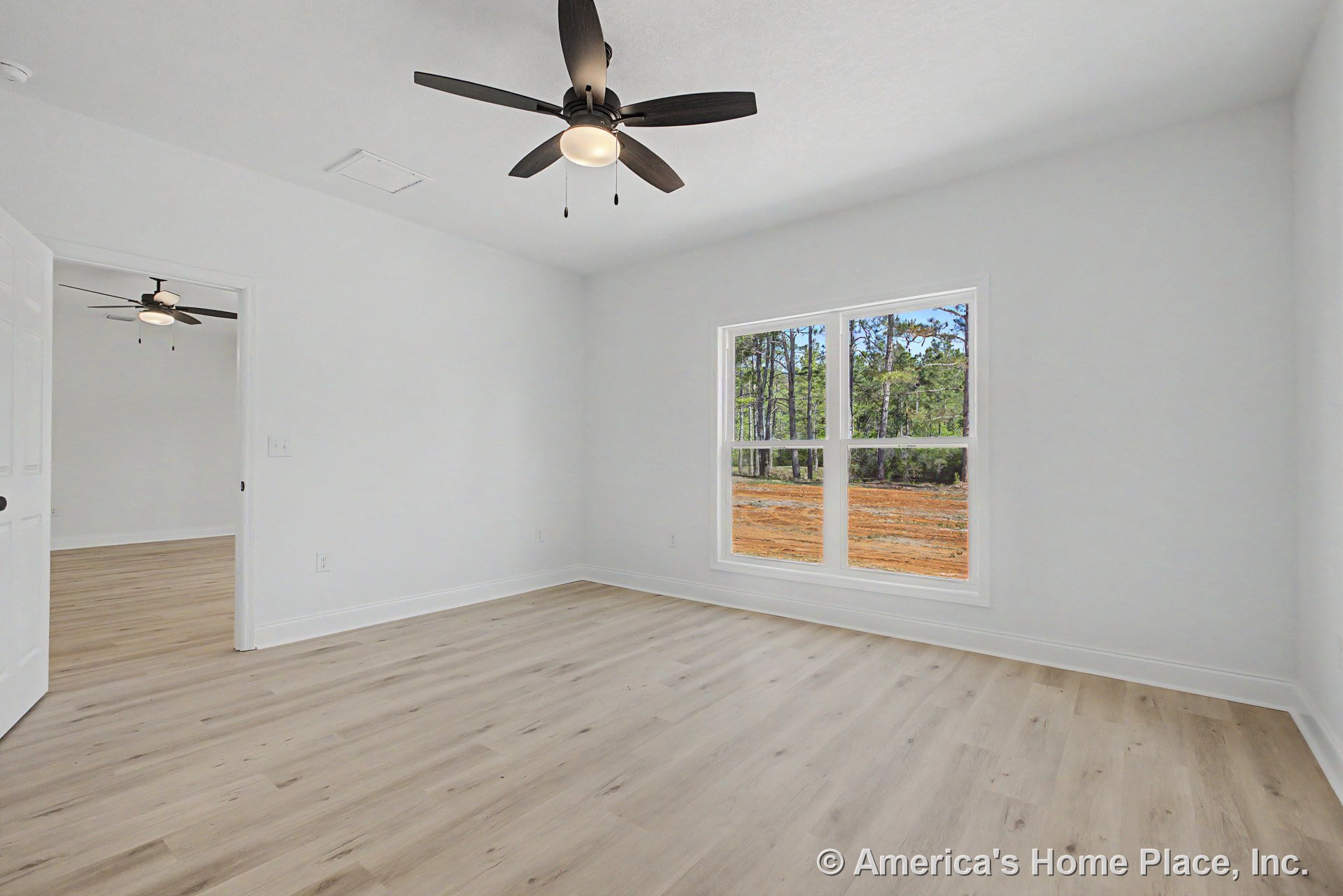 Bedroom with light wood plank flooring, large double window framed by white trim and baseboards, ceiling fan with integrated lighting on a smooth white ceiling, and open doorway