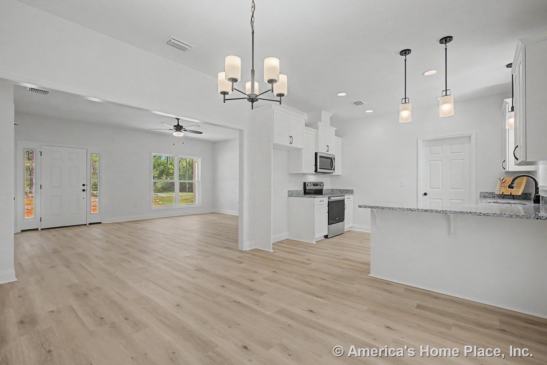 Open concept kitchen and living area with light wood plank flooring, white shaker cabinets, granite countertops, pendant and chandelier lighting, large front windows, ceiling fan