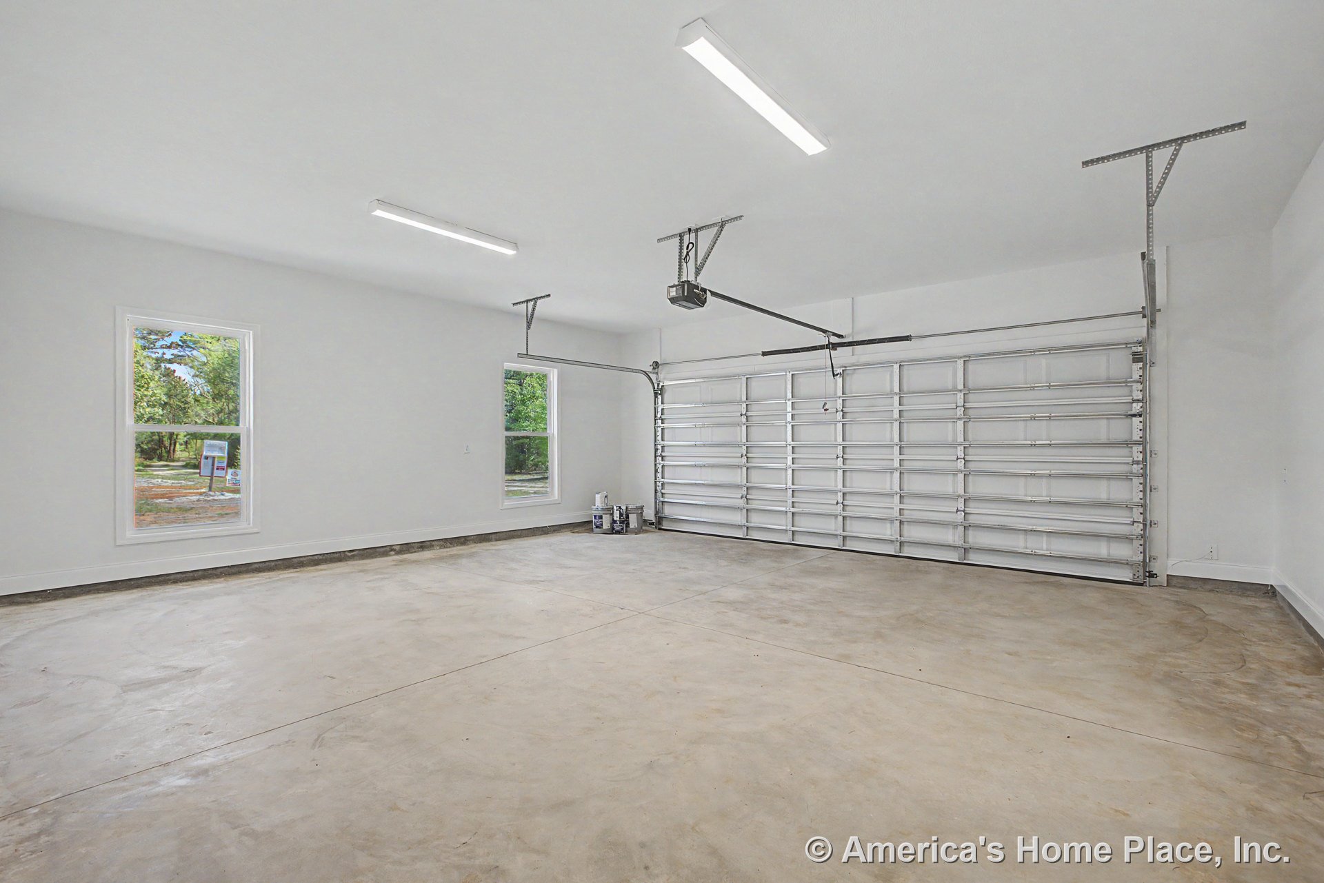 Double bay metal overhead garage door with automatic opener, concrete slab flooring, two large windows, white painted walls, ceiling-mounted fluorescent lights, and finished trim.