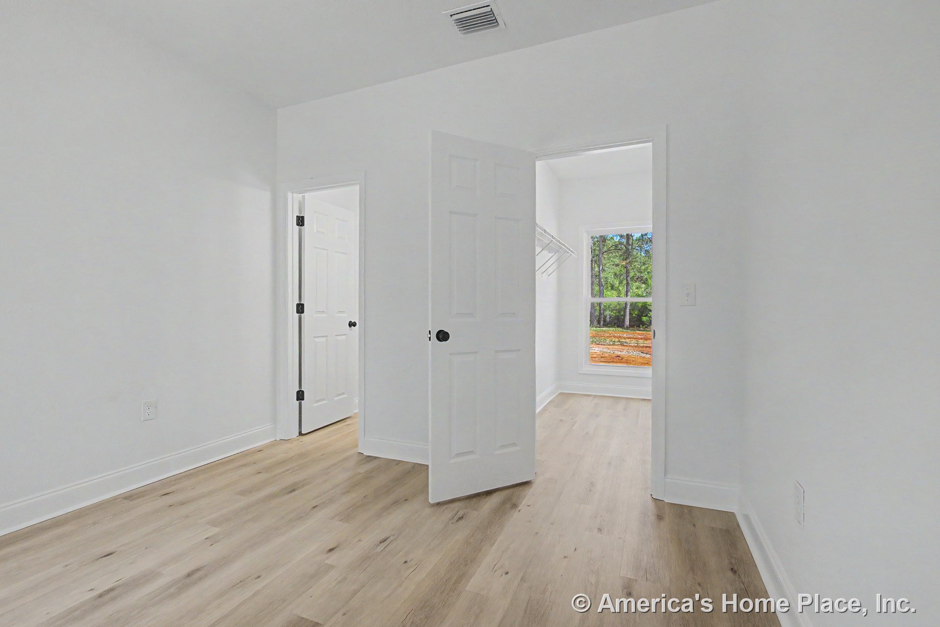 Bedroom with light wood plank flooring, white painted trim, smooth white ceiling, and white paneled doors; walk-in closet includes built-in shelving and a large window allowing