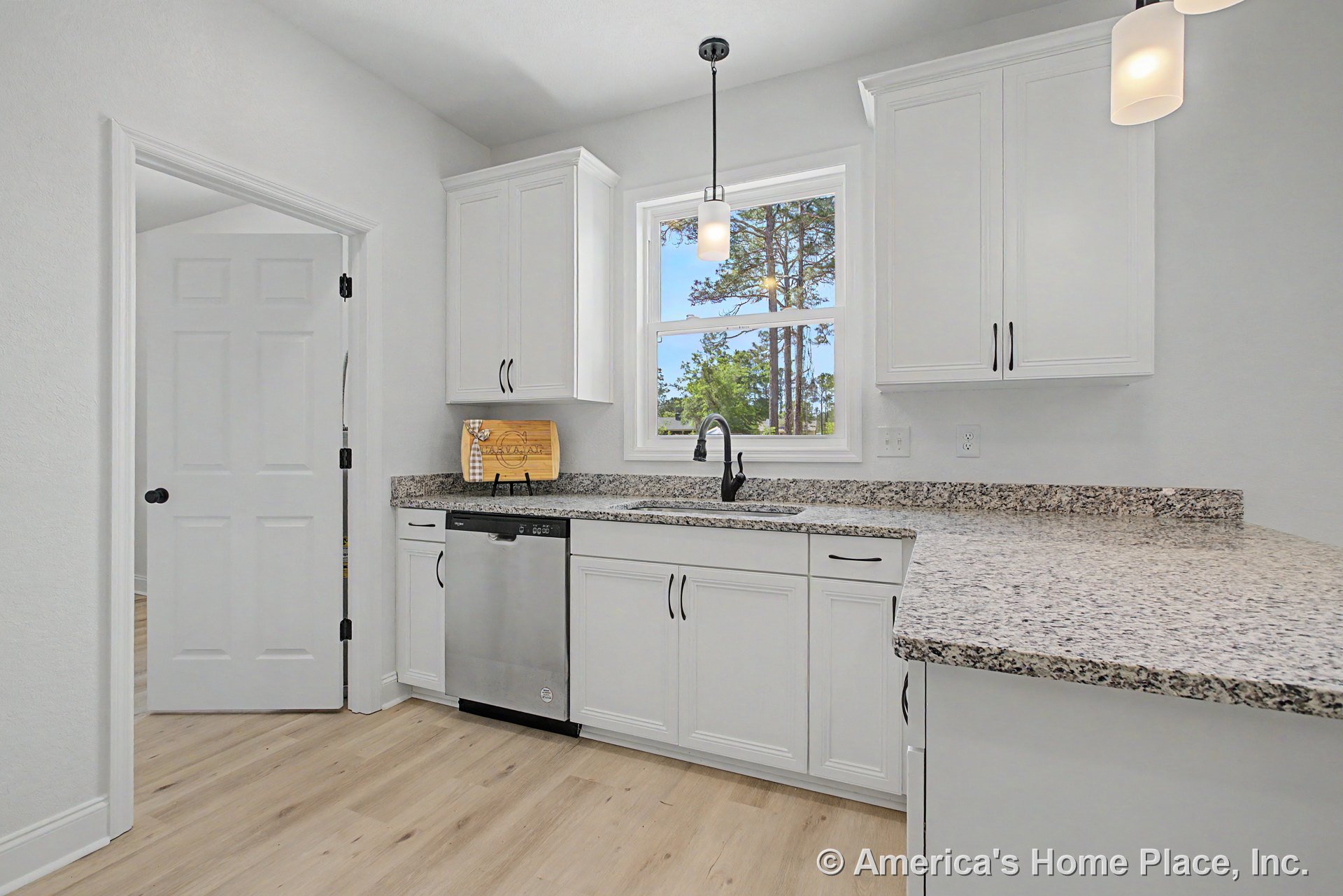 White upper and lower cabinets with brushed hardware, granite countertop and matching backsplash, stainless steel dishwasher beneath a window, single-handle black faucet, pendant