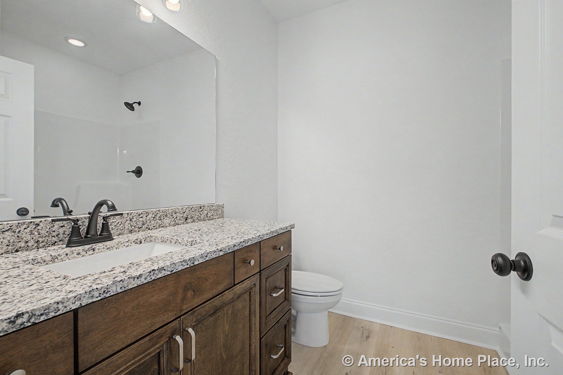 Granite vanity with undermount sink, dark wood cabinetry, matte black faucet, large wall mirror, light wood flooring, white toilet, recessed lighting, modern trim, and shower