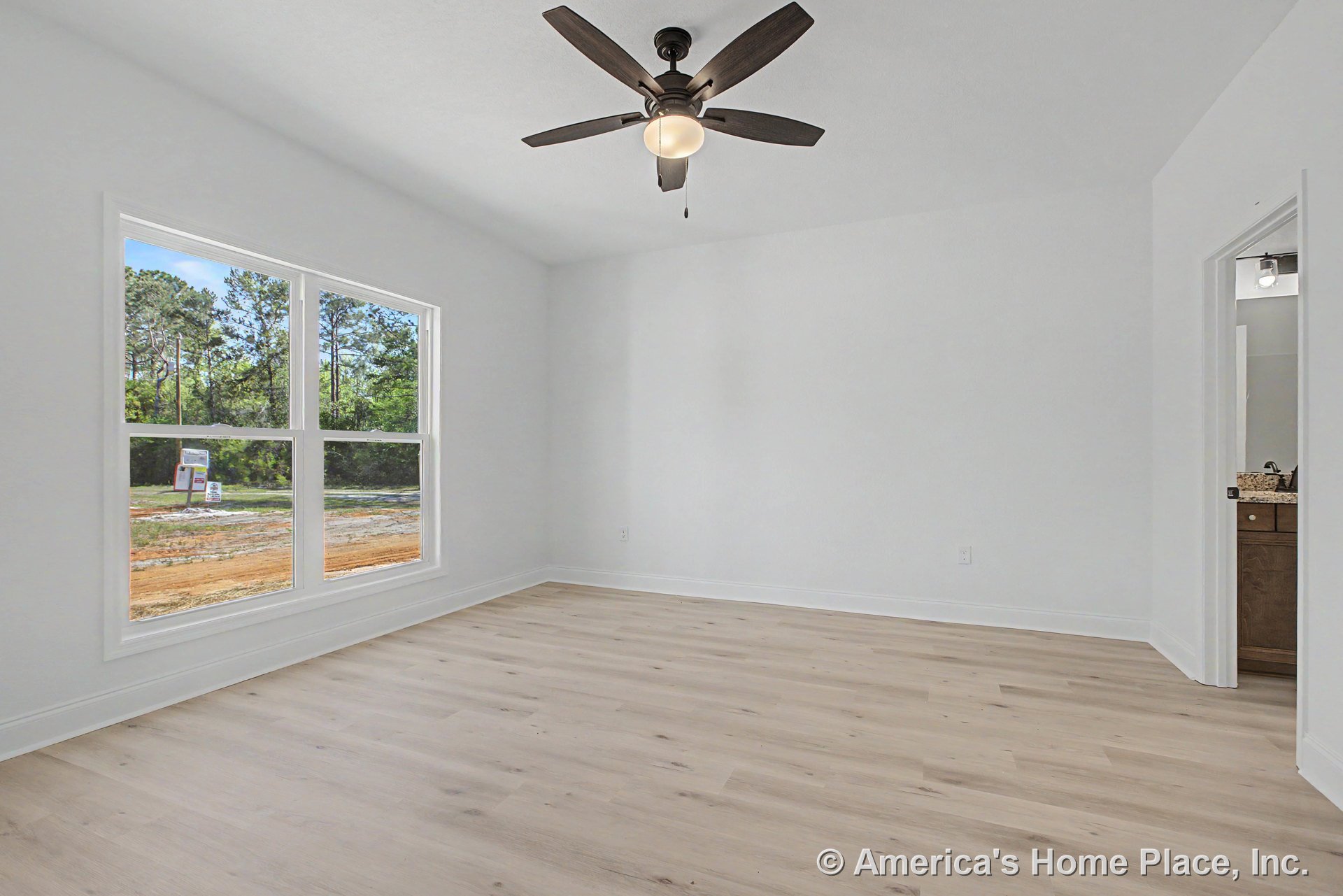 Bedroom with light wood plank flooring, large double window framed by white trim, ceiling fan with integrated light fixture, and doorway leading to bathroom with granite-topped