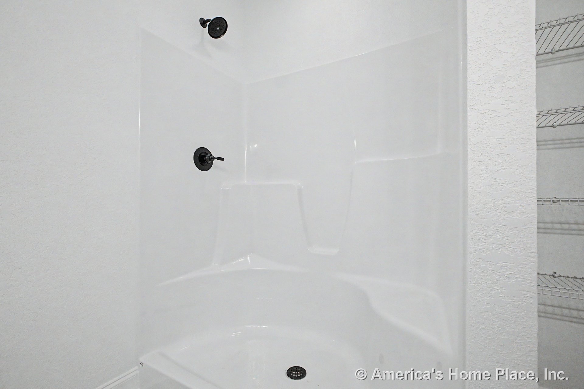 White fiberglass shower enclosure with built-in seat and shelving, matte black showerhead and faucet handle, textured white wall, wire shelving in adjacent closet.
