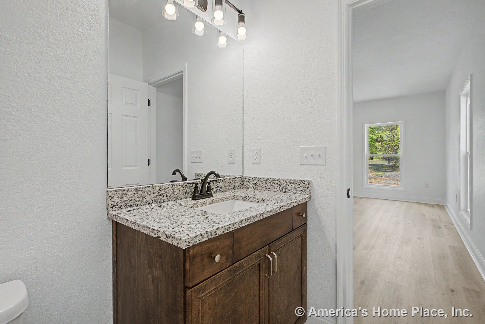 Granite bathroom vanity with undermount sink and dark bronze faucet, dark wood cabinetry, wall-mounted mirror beneath three-light fixture, white textured walls, doorway leading to