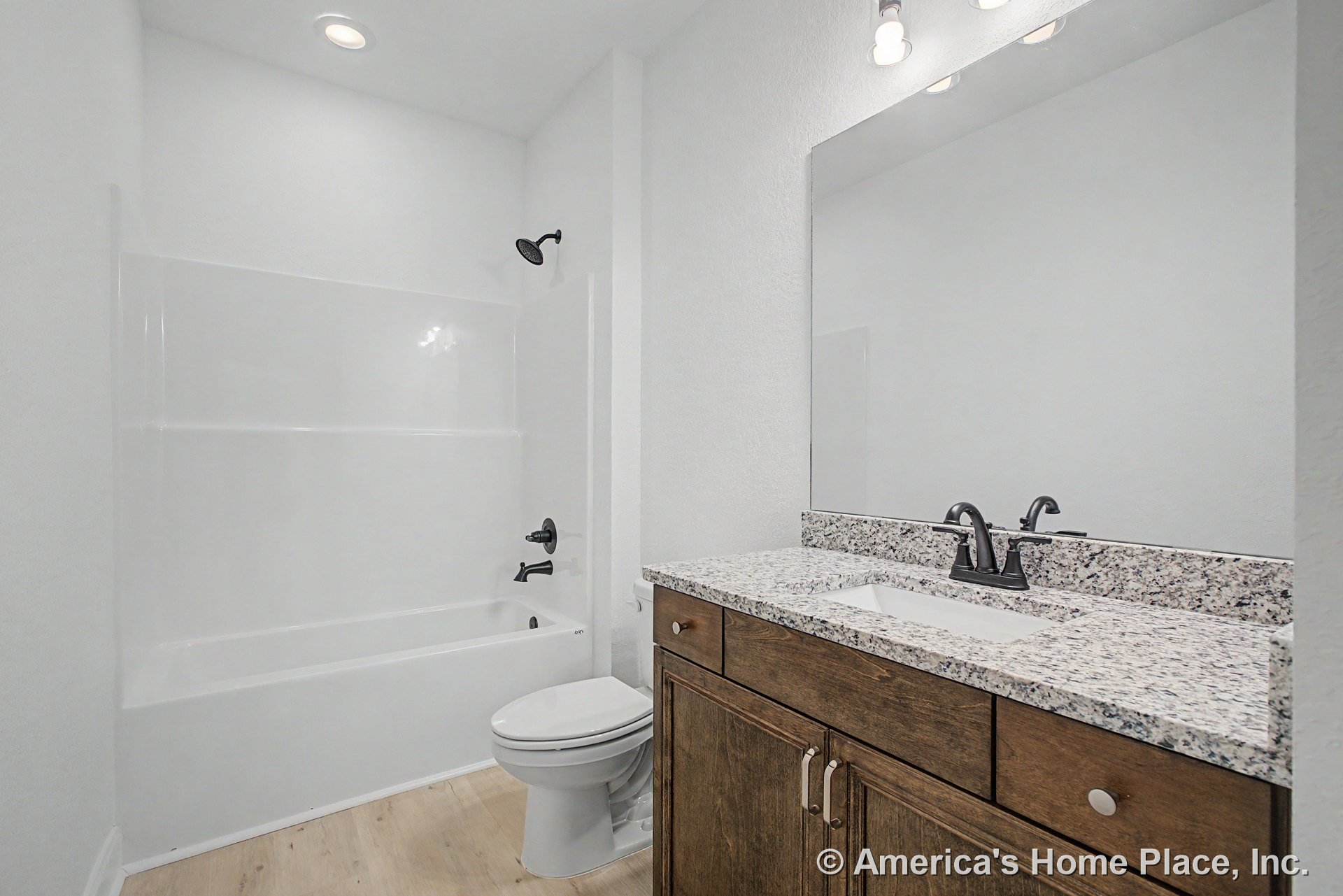 Granite countertop atop wood vanity with large wall mirror, tub-shower combination featuring black fixtures, light wood flooring, recessed lighting, and white walls.