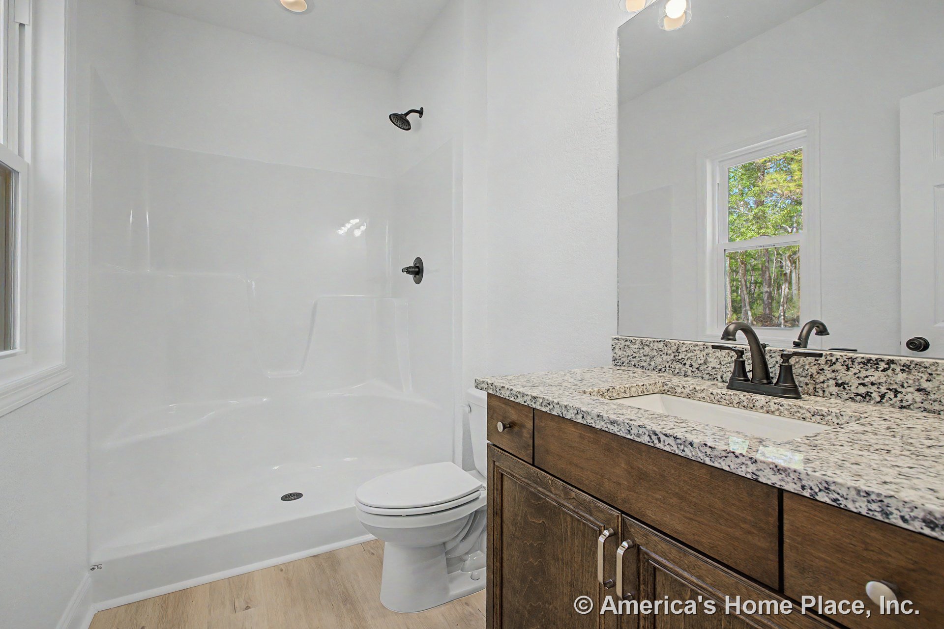 Single vanity with granite countertop and dark wood cabinetry, fiberglass shower enclosure, large wall mirror, single lever faucet, light wood plank flooring, white walls, built-in