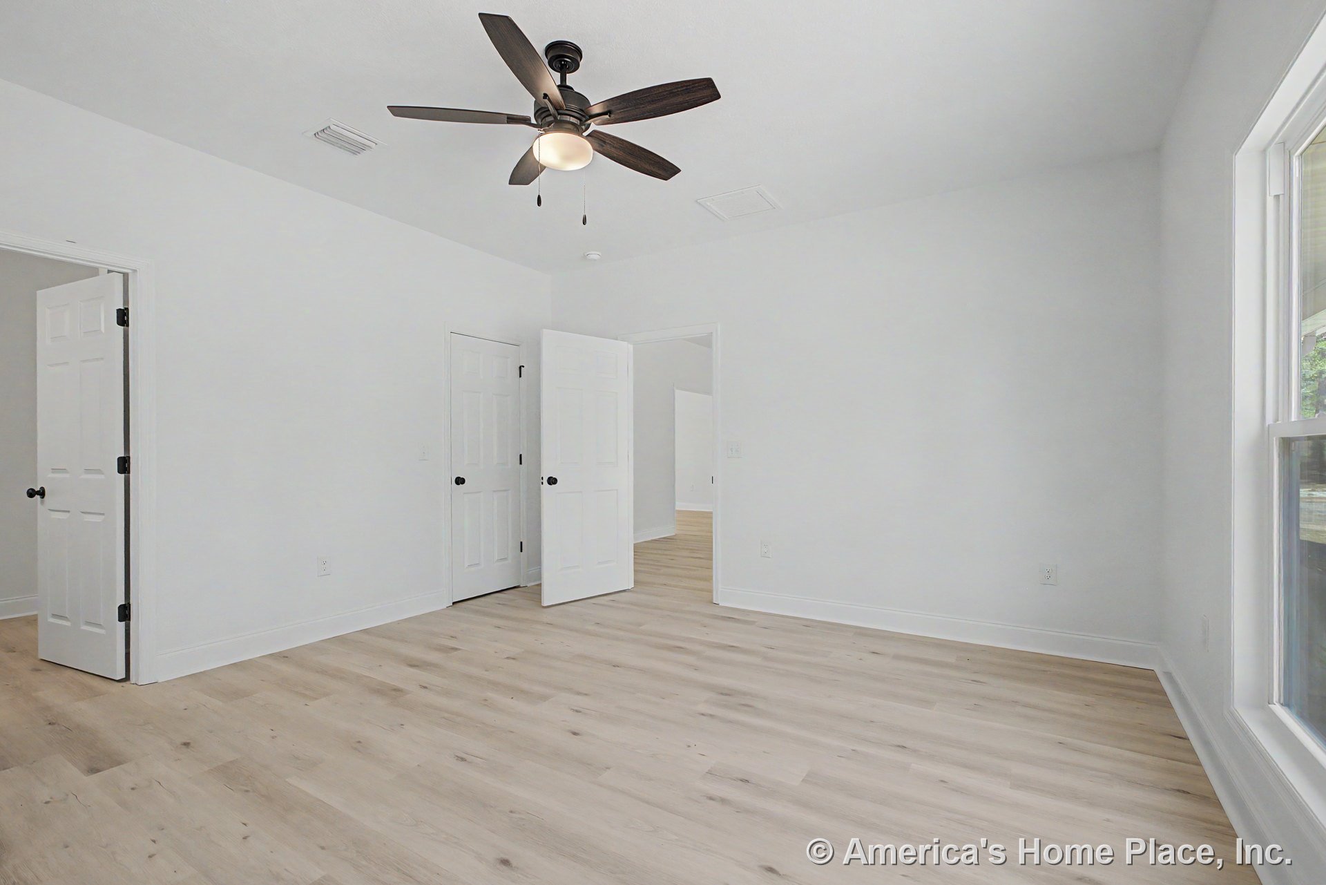 Bedroom with light wood plank flooring, white walls, smooth white ceiling, ceiling fan with integrated lighting, large window with white trim, white interior doors, and white