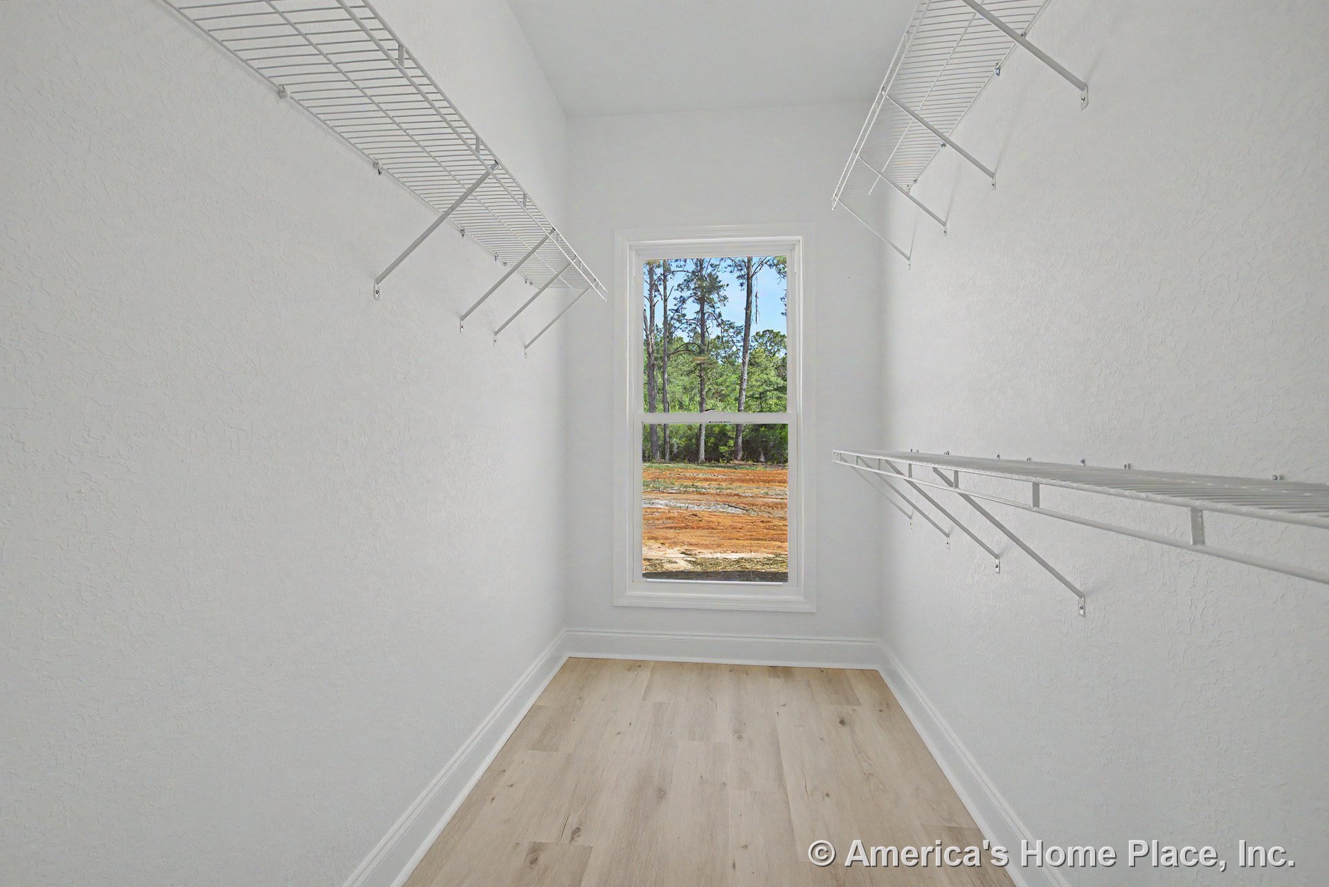 Walk-in closet with white wire shelving, large window framed with trim, light wood flooring, textured white walls, baseboard molding, and high ceiling.